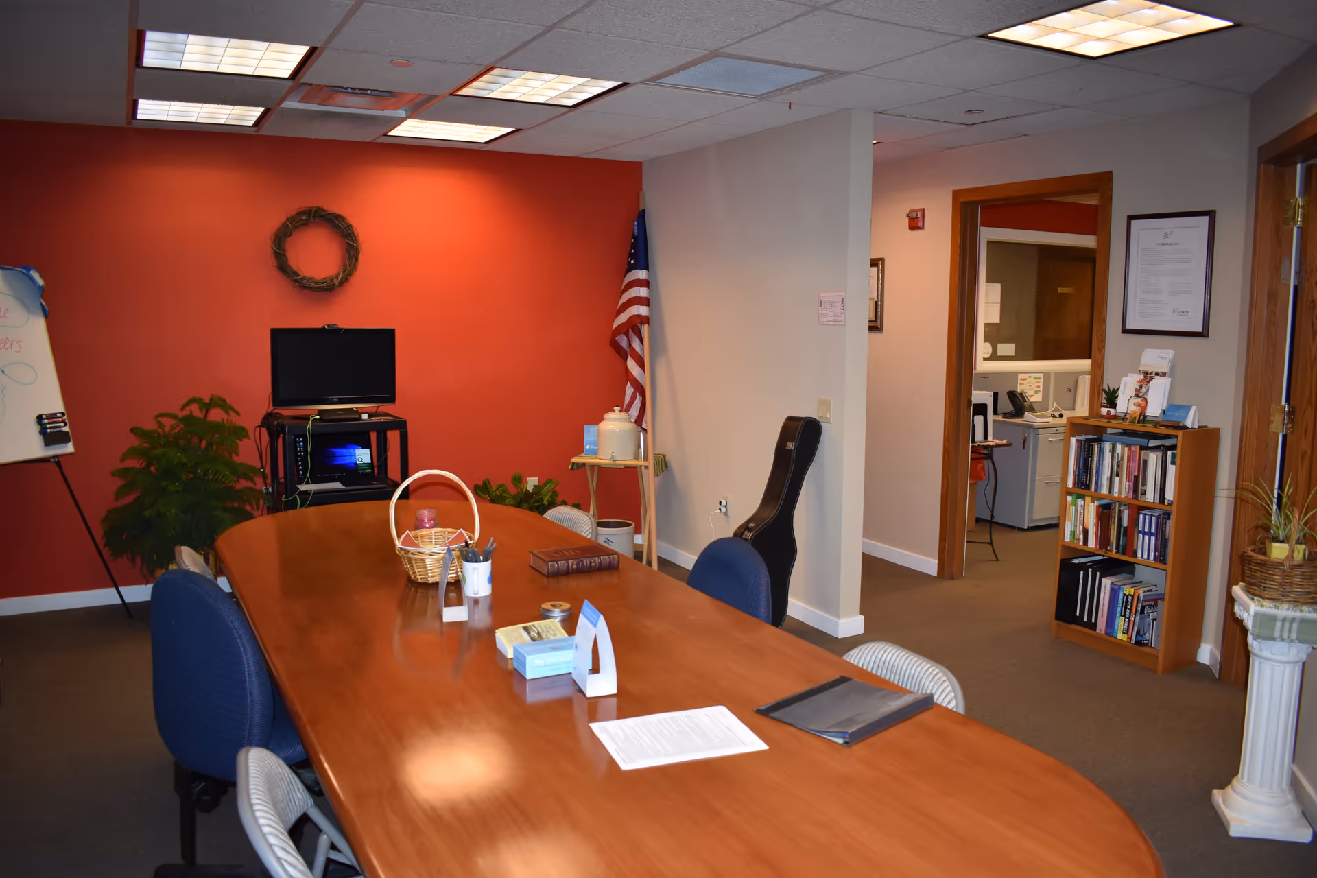 A conference room with a long wooden table, chairs, red accent wall, and bookshelf.