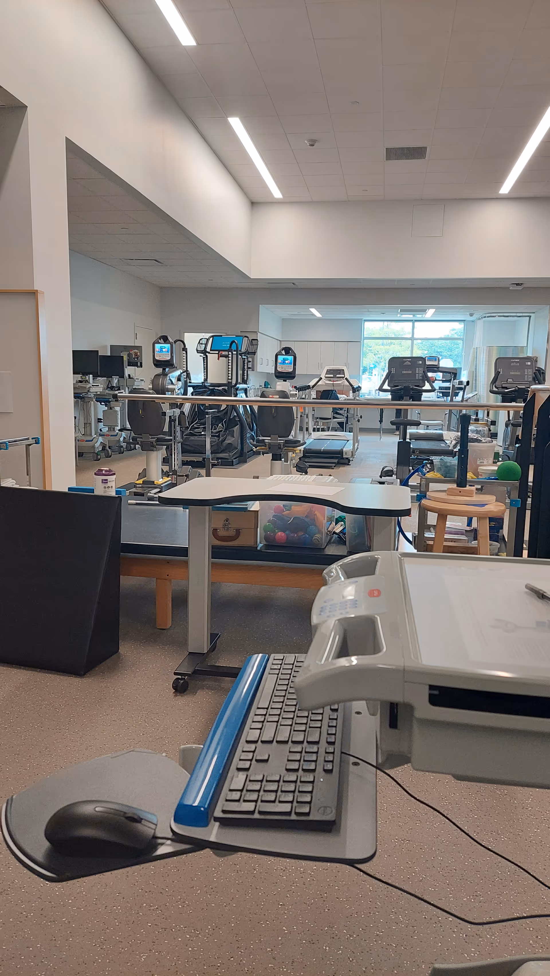 Indoor rehabilitation gym with cardio machines and a desk in the foreground.