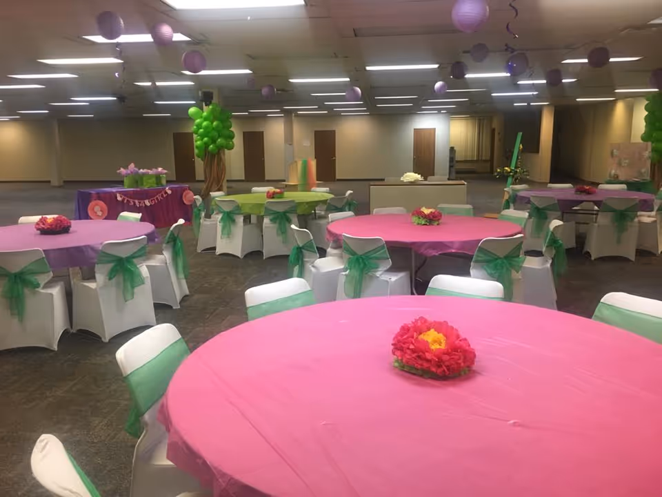 Decorated indoor banquet hall with pink tables, white chairs, green sashes, and balloons.