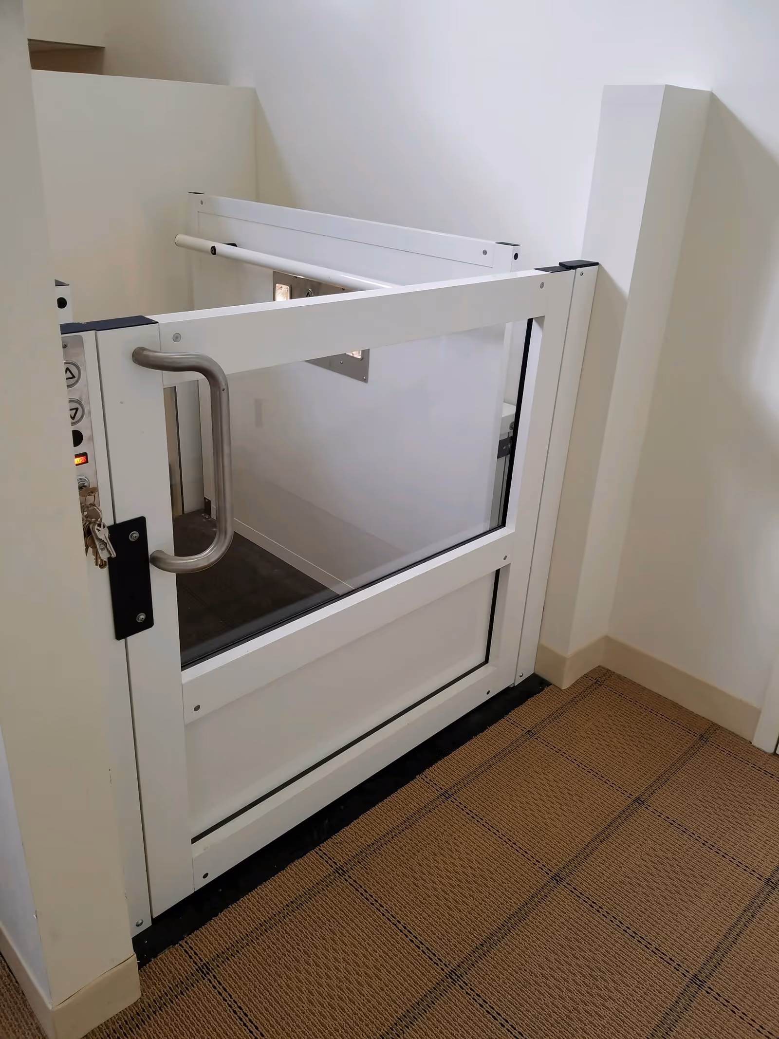 White accessibility lift with glass panels in a hallway corner.