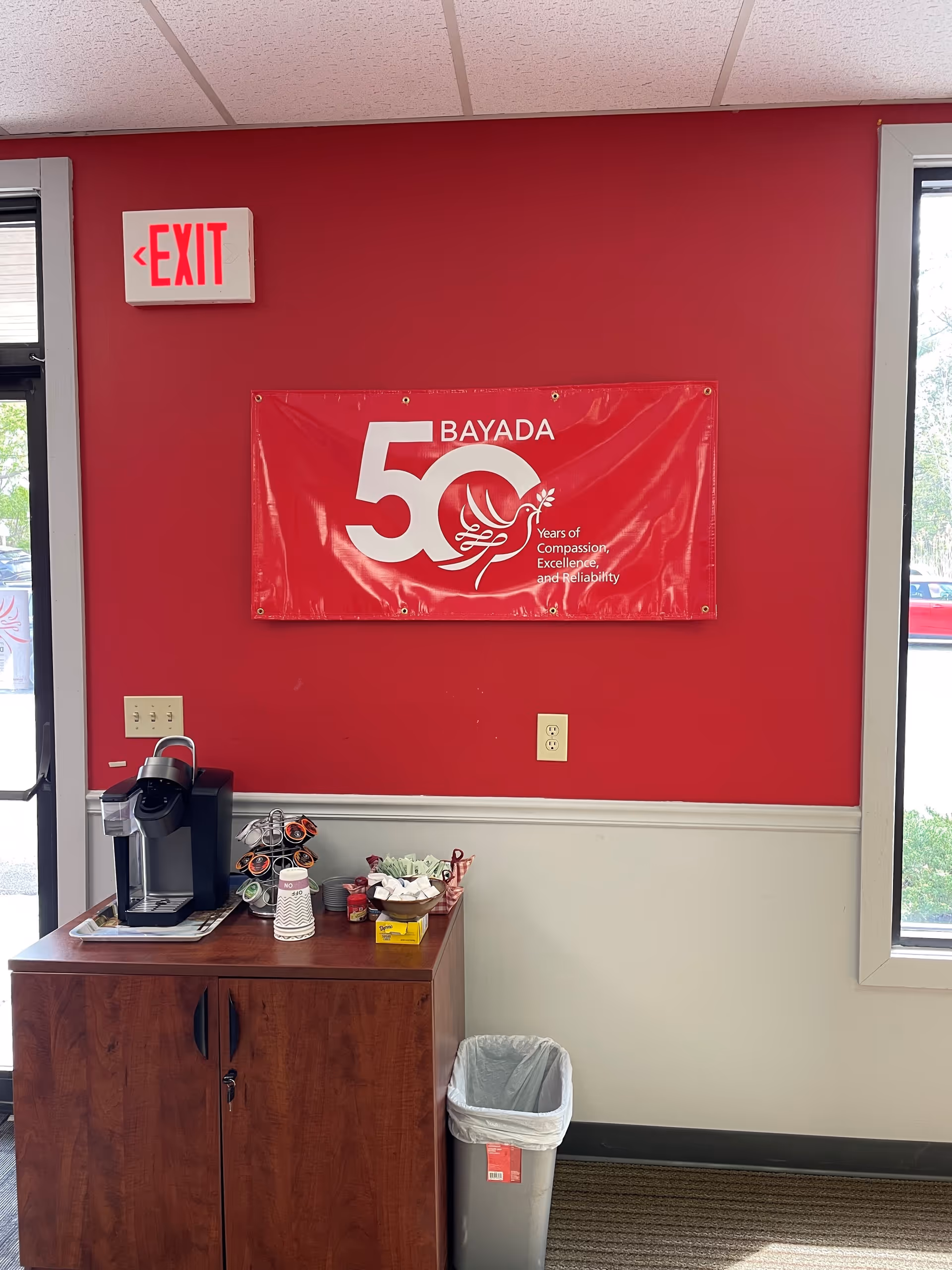 Red-walled interior featuring a coffee station, BAYADA banner, exit sign, and trash can.