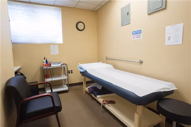 A treatment table in a small beige room with a clock, window, and medical cart.