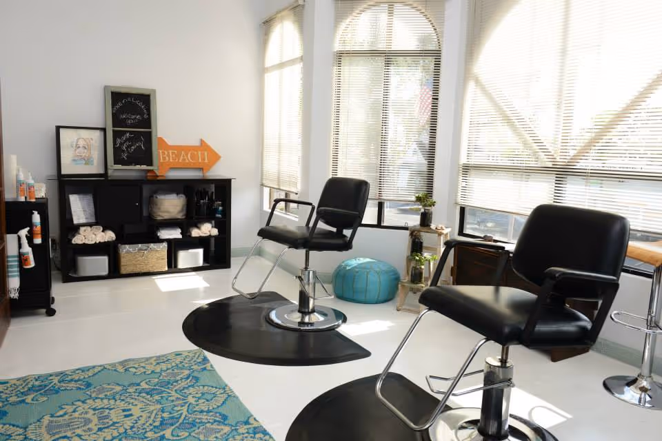 Interior salon area with black styling chairs, shelves, and large windows letting in light.