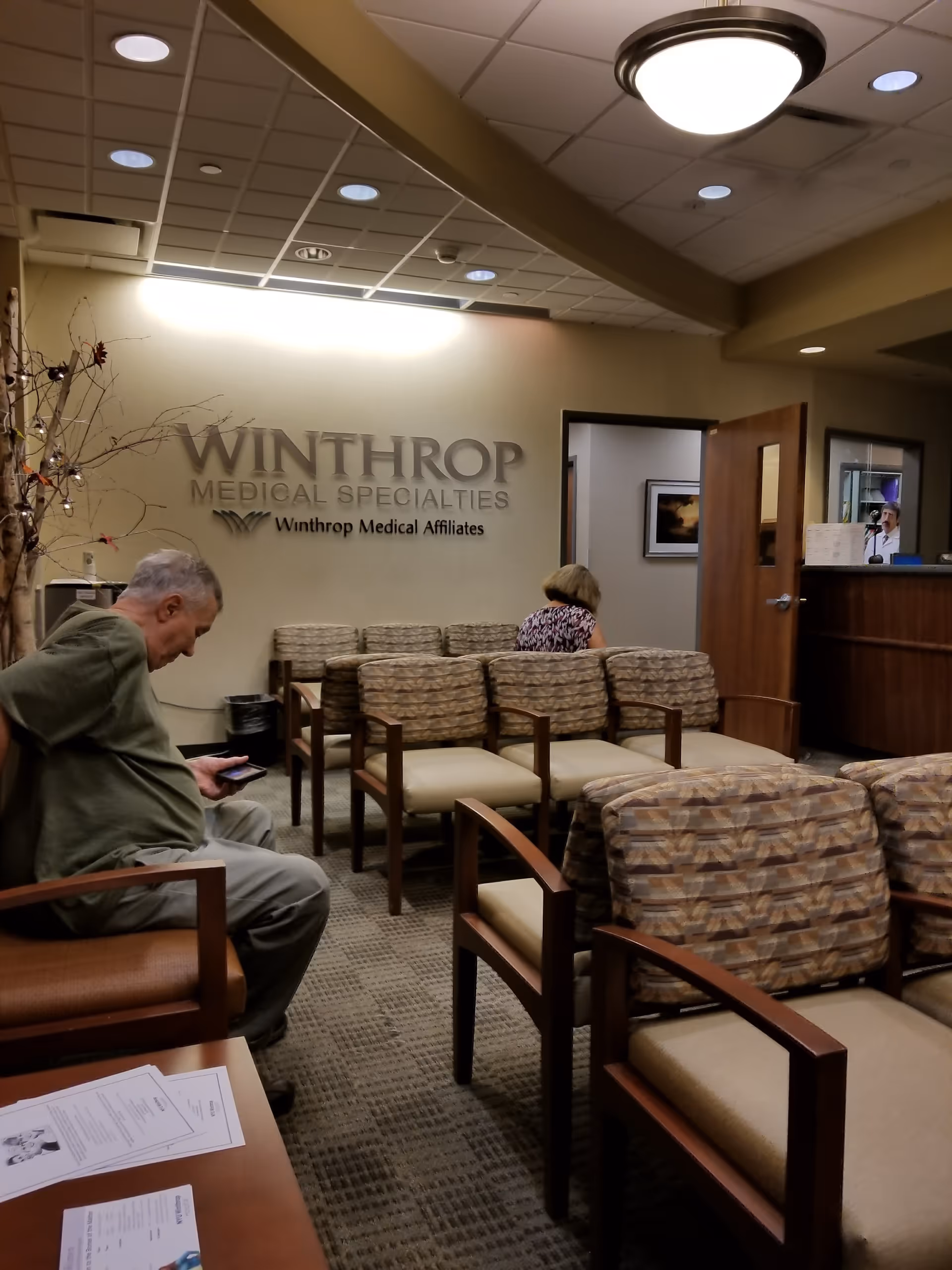 A medical office waiting room with beige chairs, a reception desk, and two people seated.