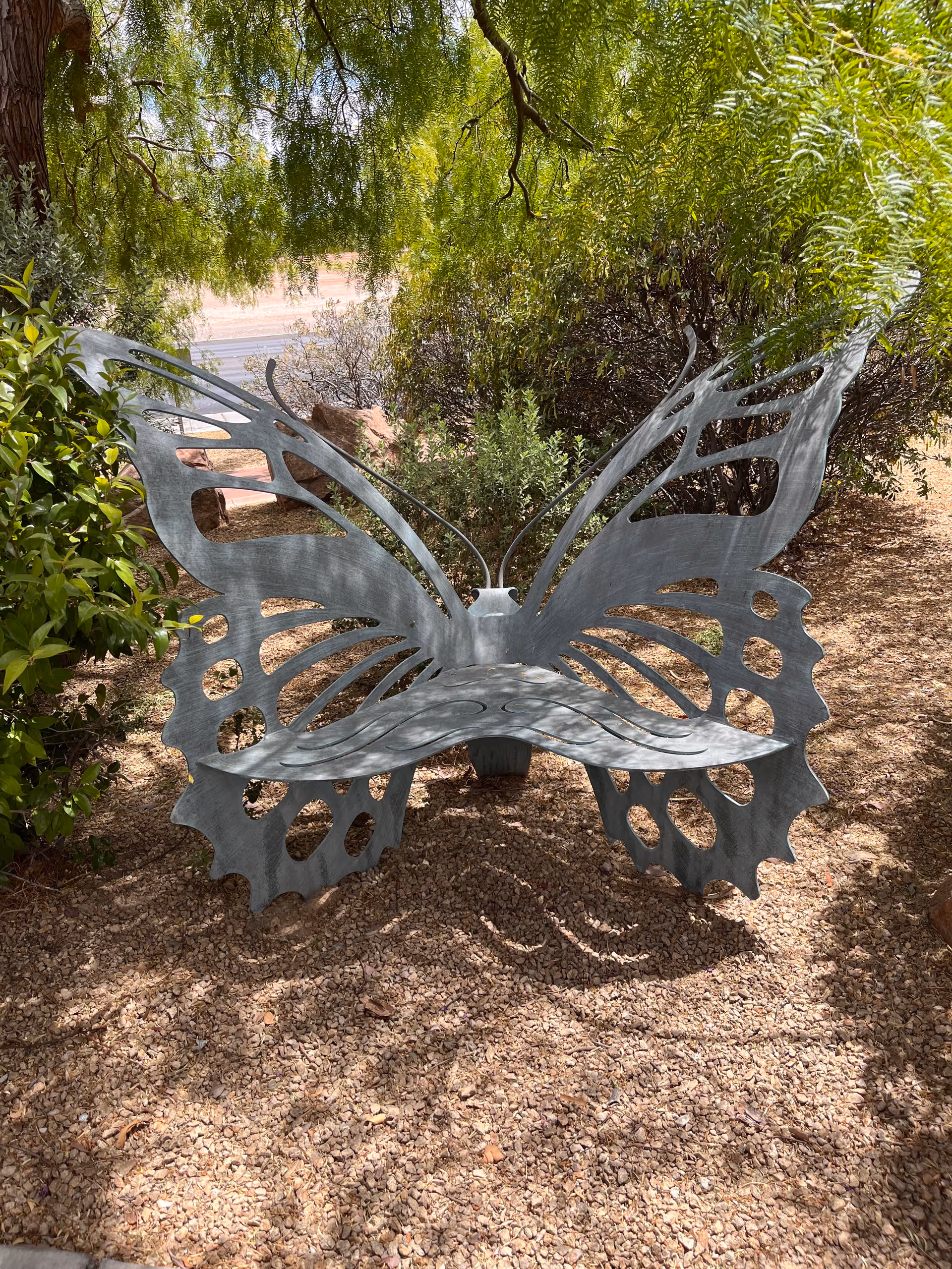 Metal butterfly-shaped bench sculpture in a sunny outdoor garden with gravel ground.