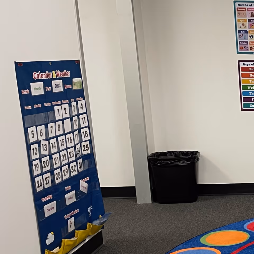 Blue calendar board with dates and a trash can in a bright hallway corner.