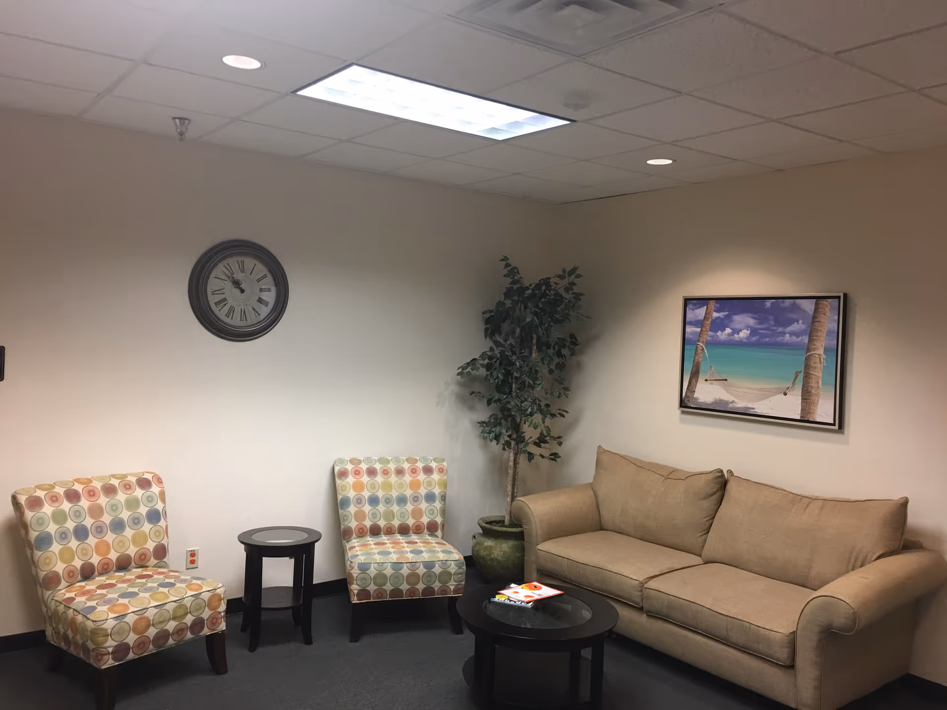 Lounge seating area with beige sofa, colorful chairs, plant, clock, and beach artwork.