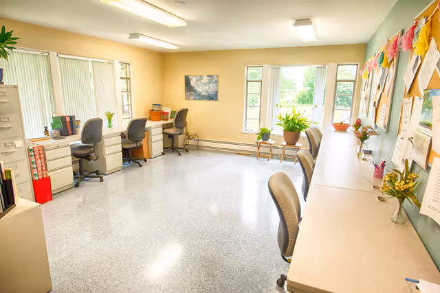 Bright interior office area with desks, chairs, bulletin boards, and windows.
