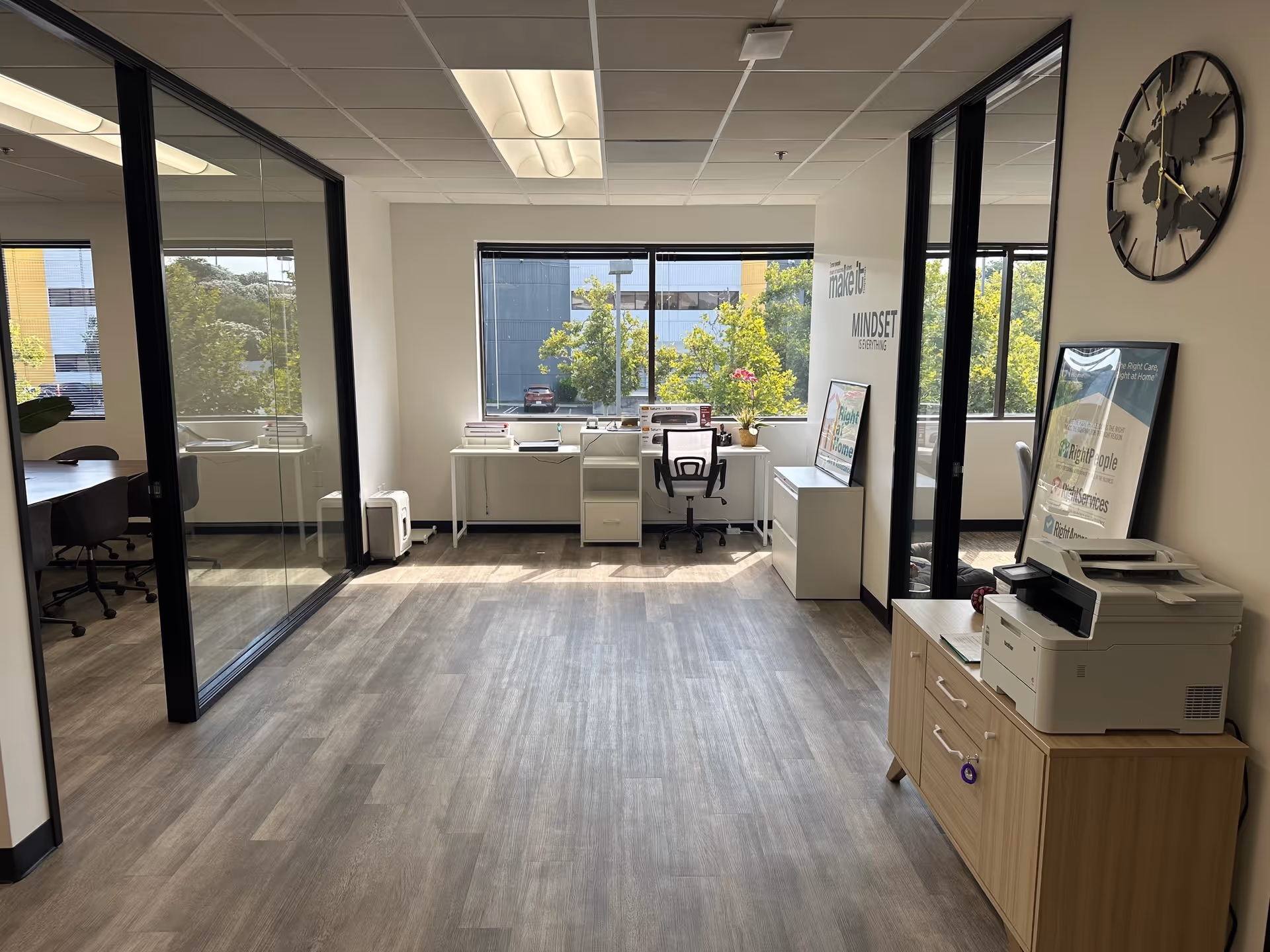 Modern office interior with glass partitions, desks, and a printer by large windows.