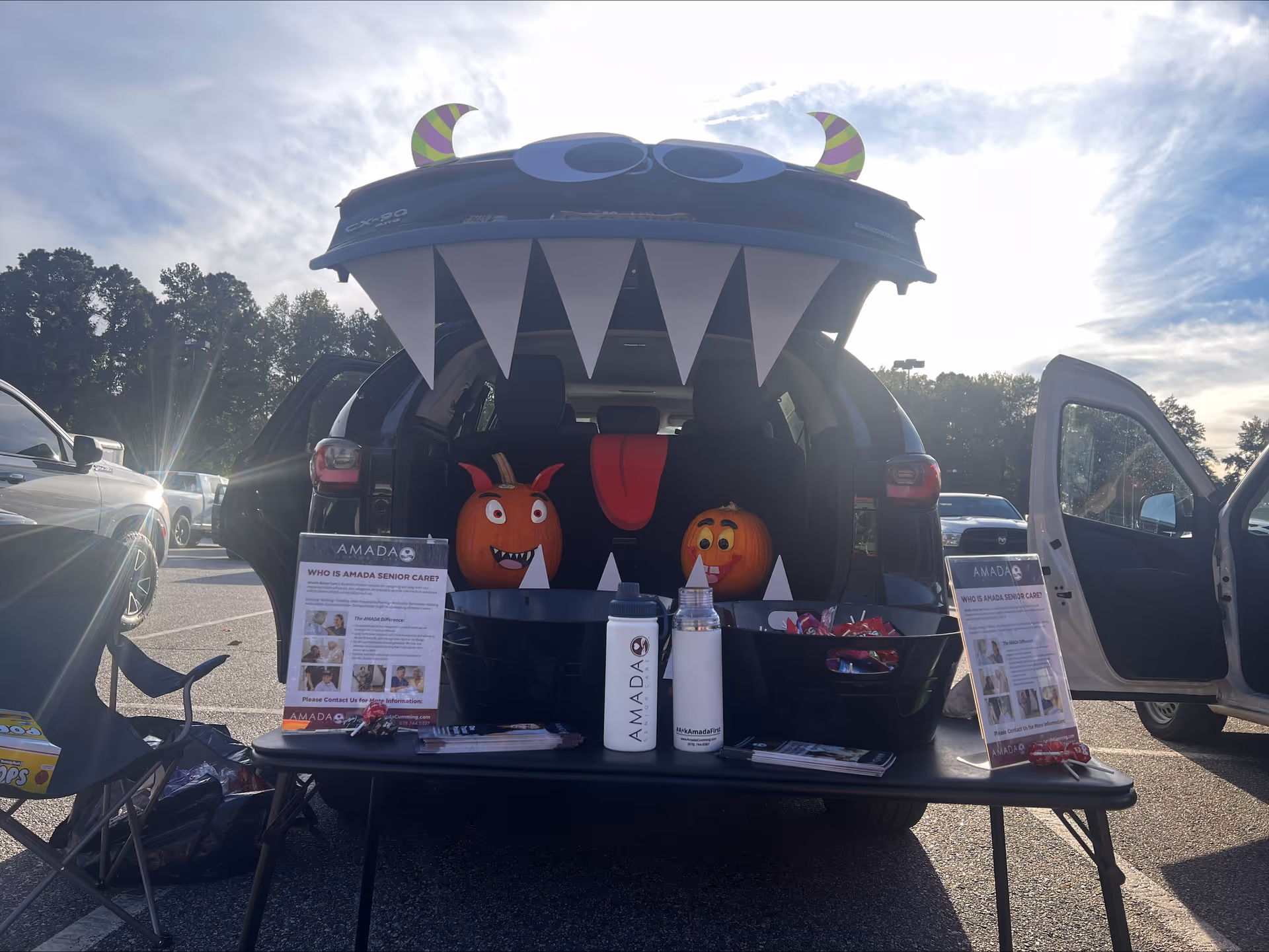 Car trunk decorated as a monster with pumpkins, flyers on a table in a parking lot.
