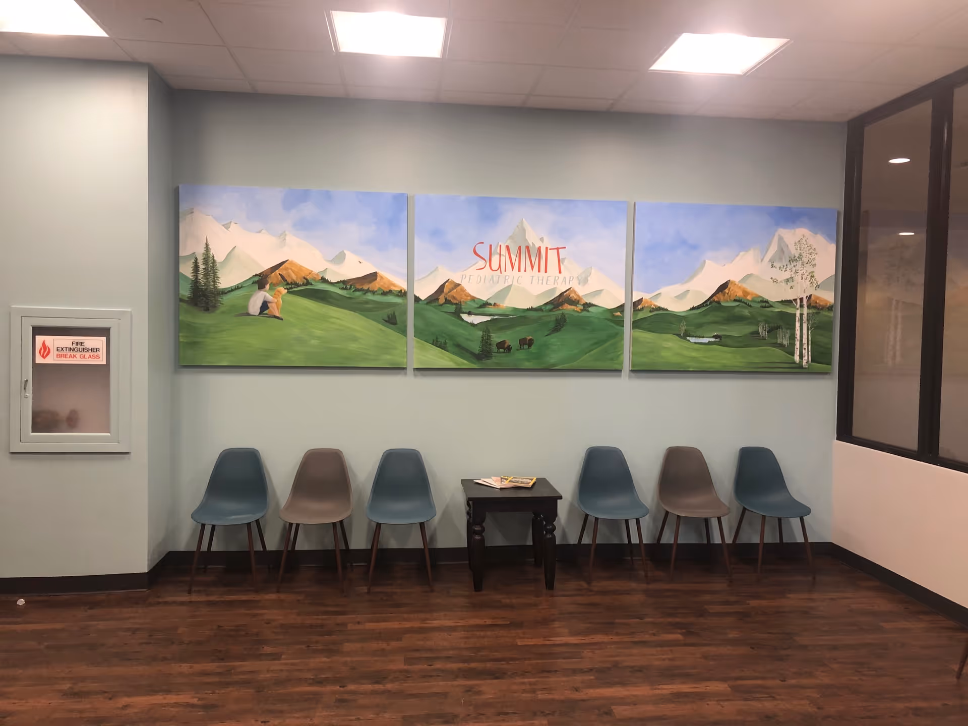 Row of chairs along a pale blue wall with a three panel mountain mural and magazines.