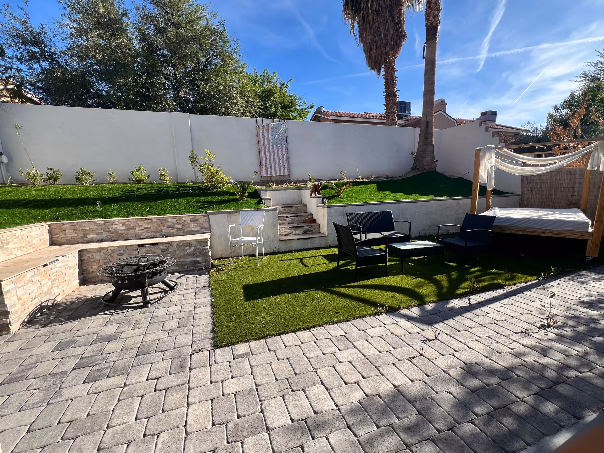 Sunlit backyard at Amore Healthcare featuring a paved patio, fire pit, seating, and canopy bed.