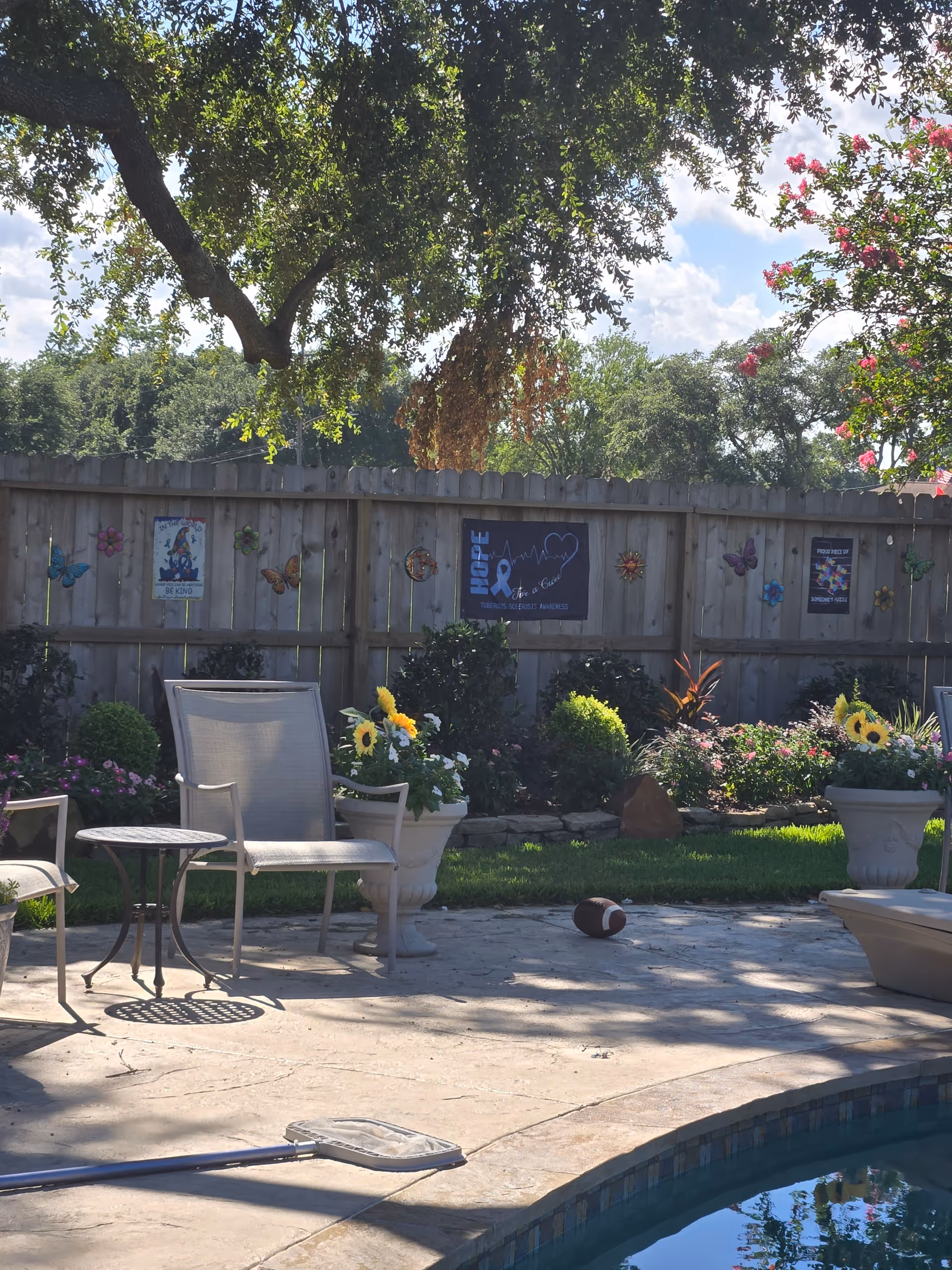 Sunny outdoor patio with chairs, flowers, and a pool beside a wooden fence.