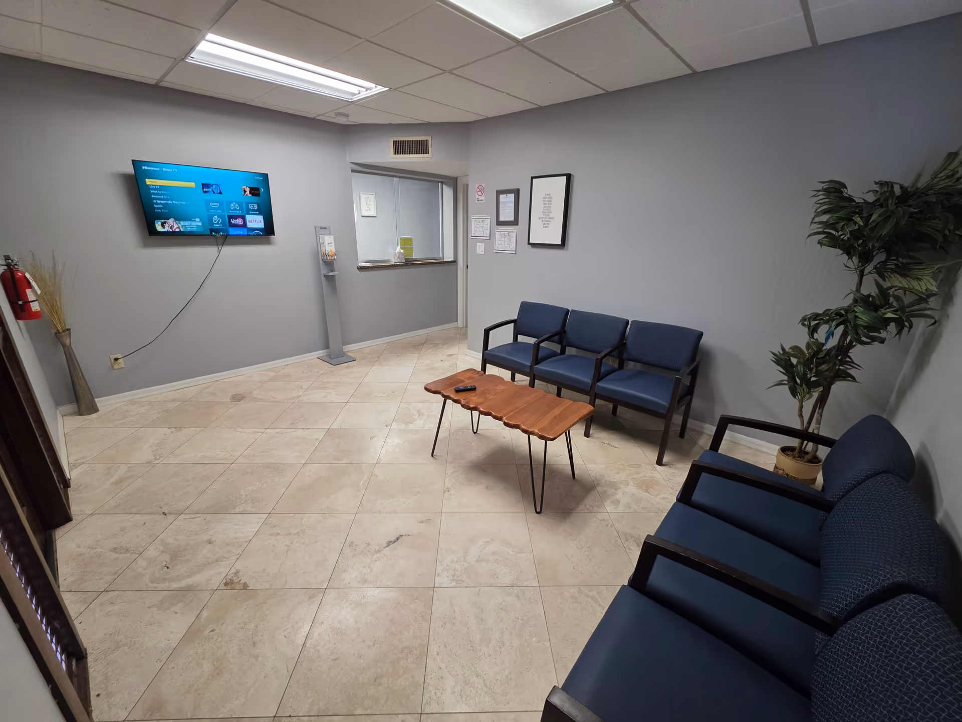 Interior waiting area with blue chairs, a wooden coffee table, TV, and plant.