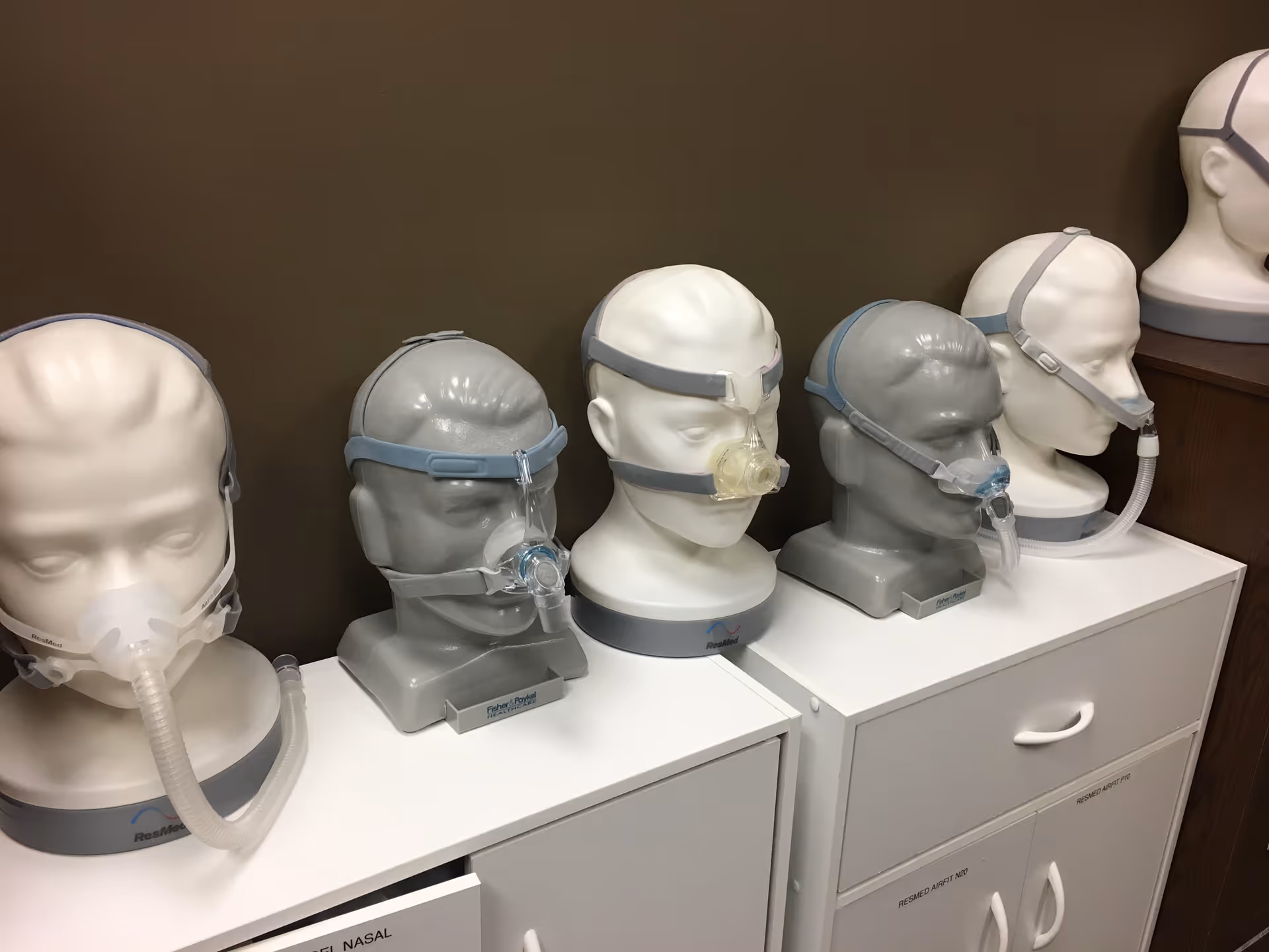 Row of CPAP mannequin heads with masks atop white cabinets in a medical equipment room.