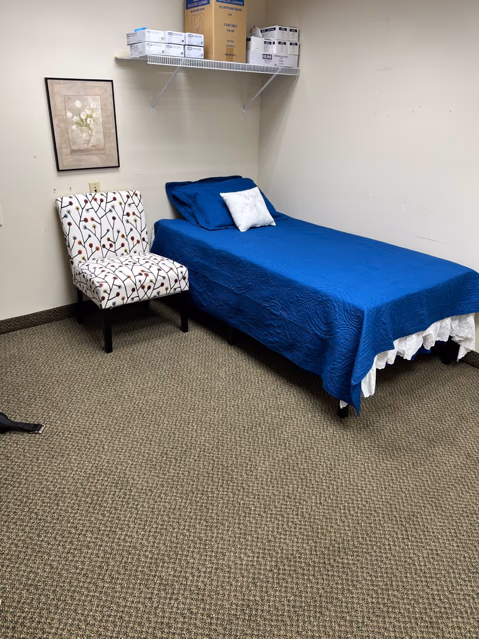 A small bedroom with a blue bedspread, patterned chair, and wall art in a storage room.