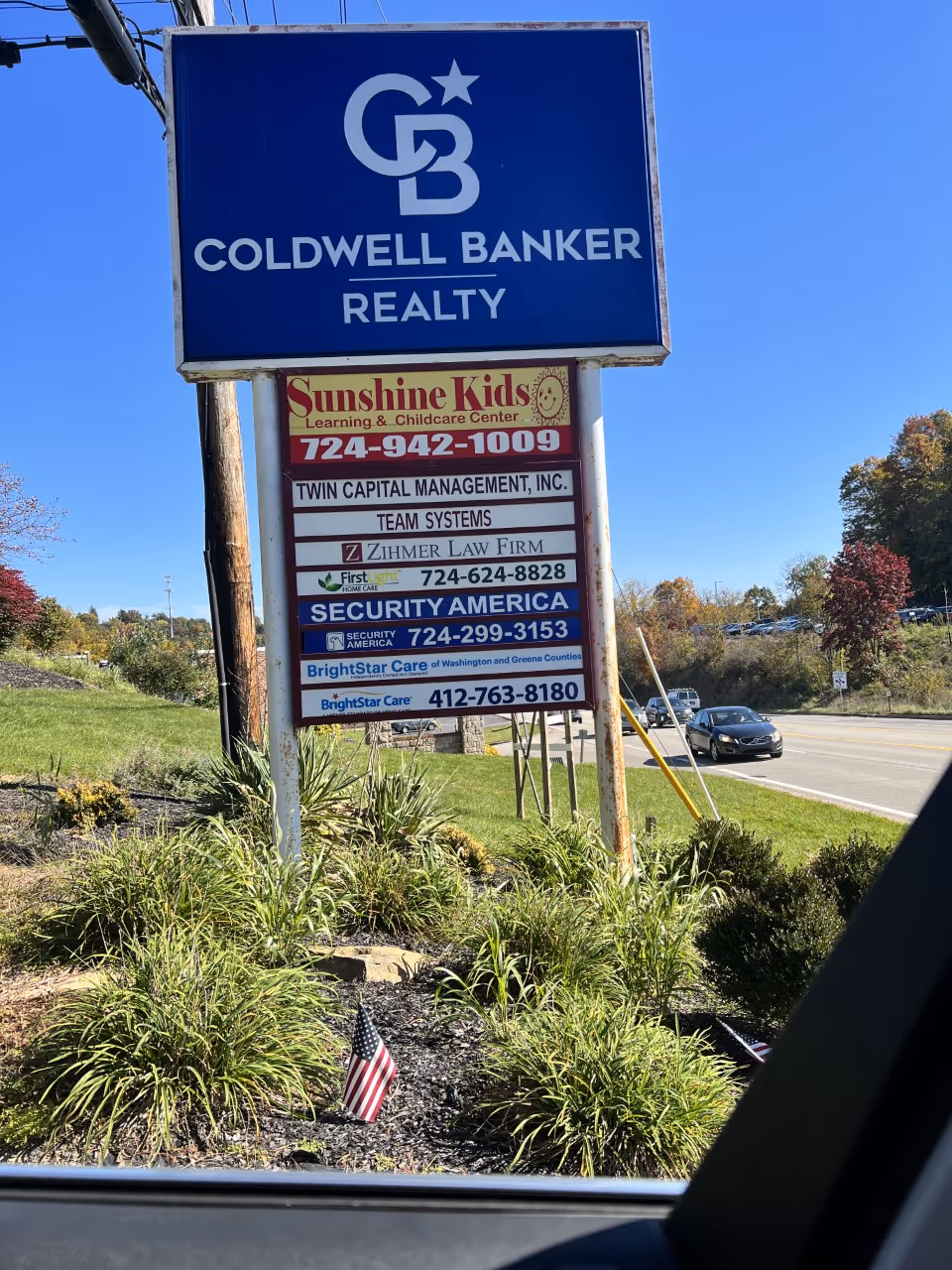 Tall blue sign for Coldwell Banker Realty with smaller business signs and landscaping along the road.