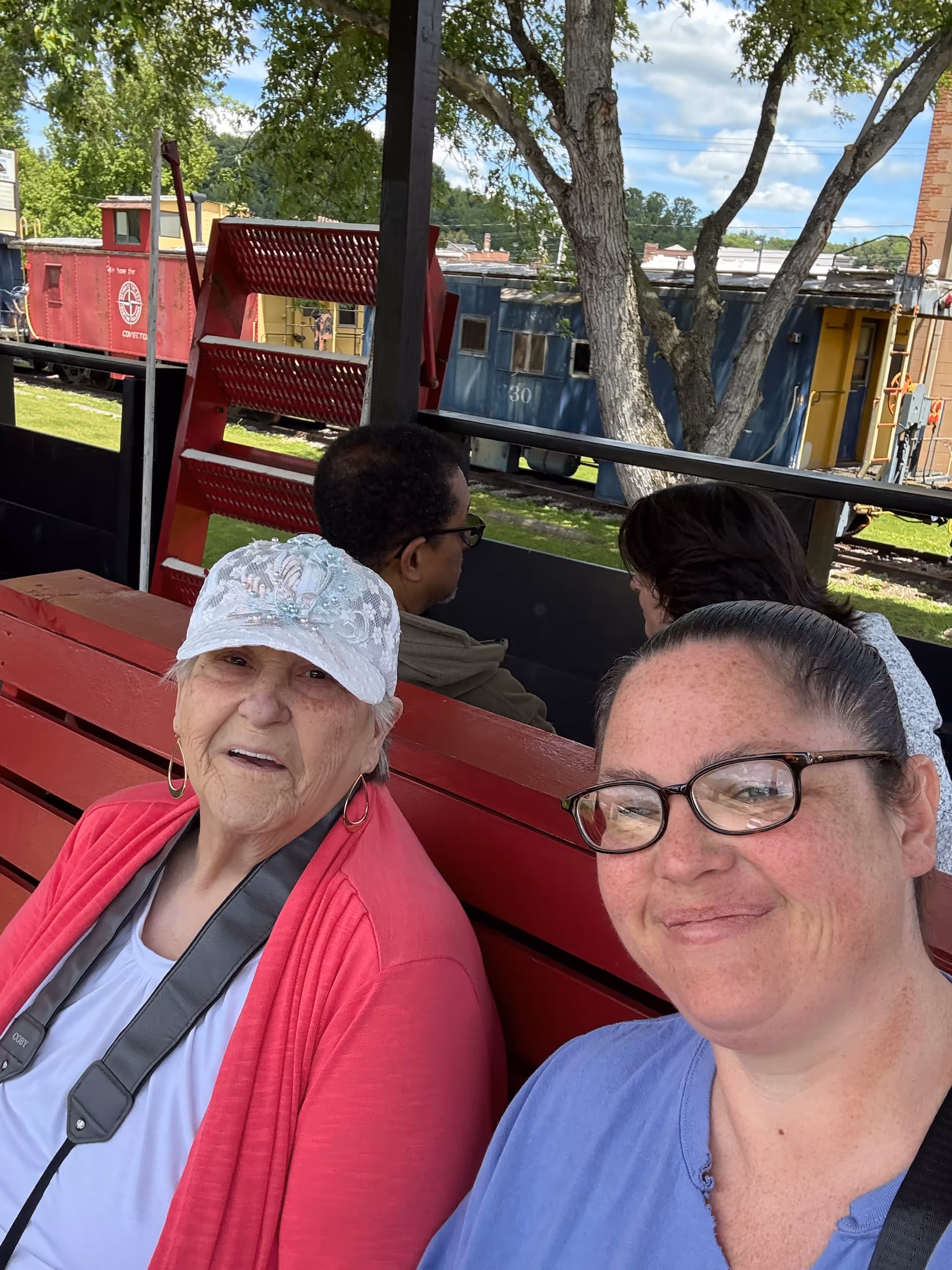 Two women sit on a red bench outdoors with trees and train cars behind.