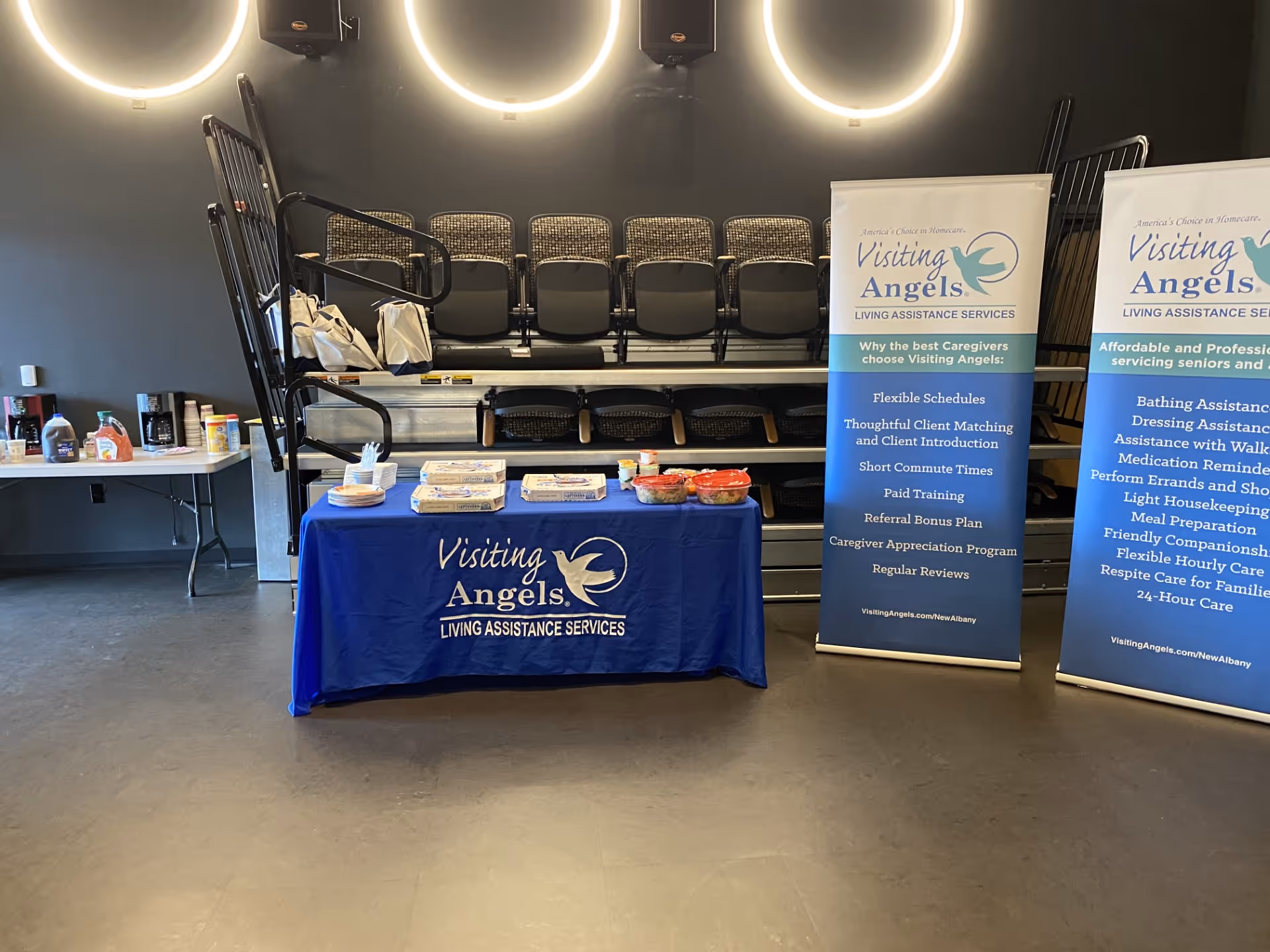 Indoor event booth with a blue Visiting Angels table, banners, and stacked chairs.