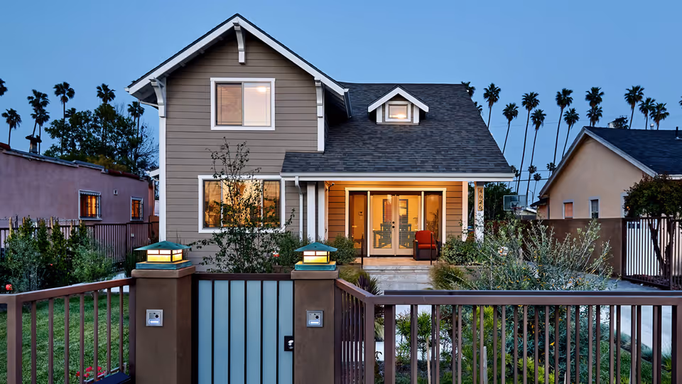 Two-story house with a gated front yard, porch seating, and palm trees in the background