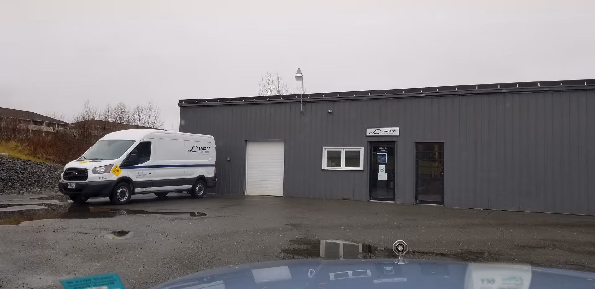 Grey metal building exterior with a LINCARE sign, a parked van, and a wet parking area.