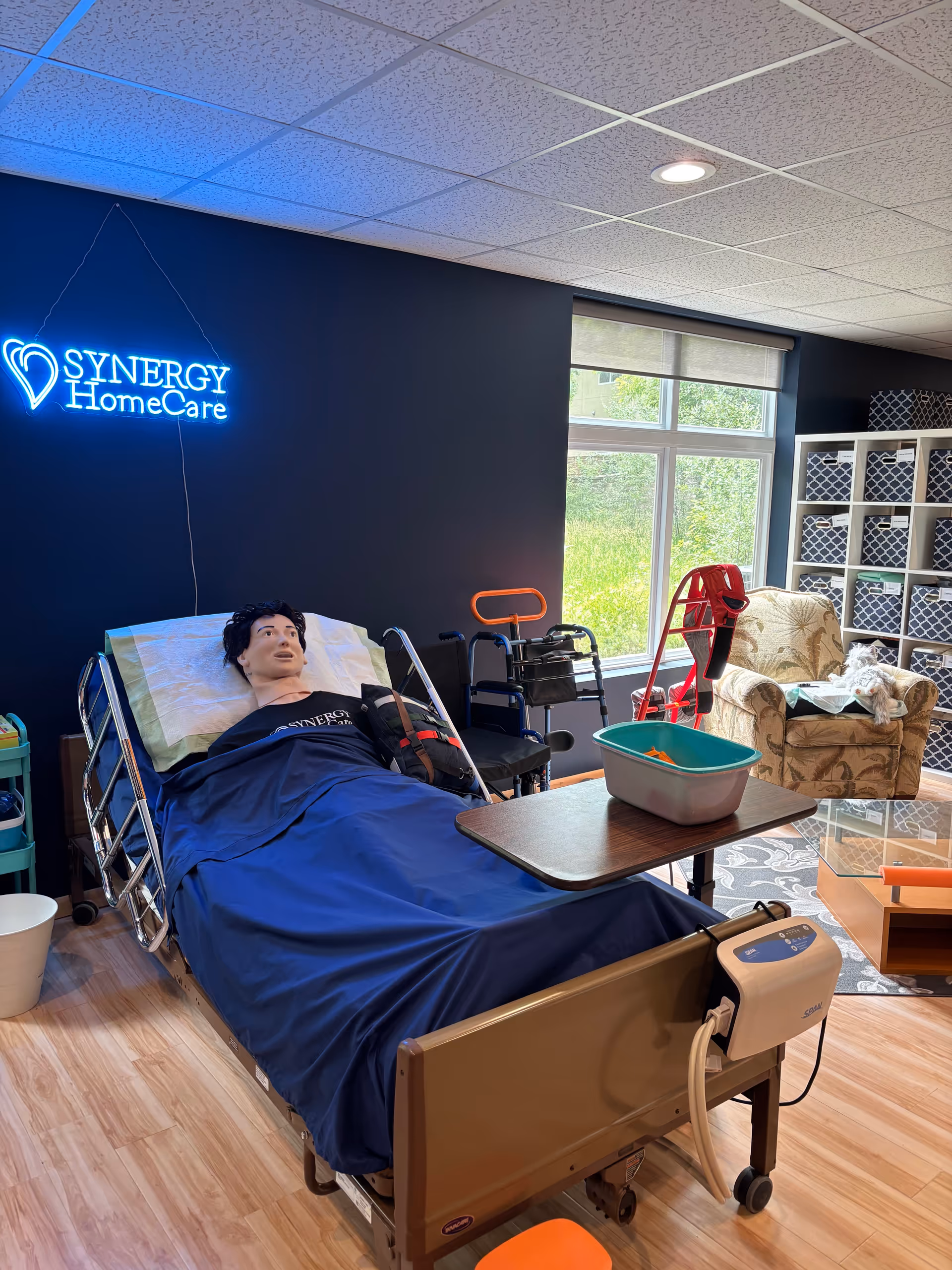 A hospital-style bed in a care room with a SYNERGY HomeCare neon sign and medical equipment.
