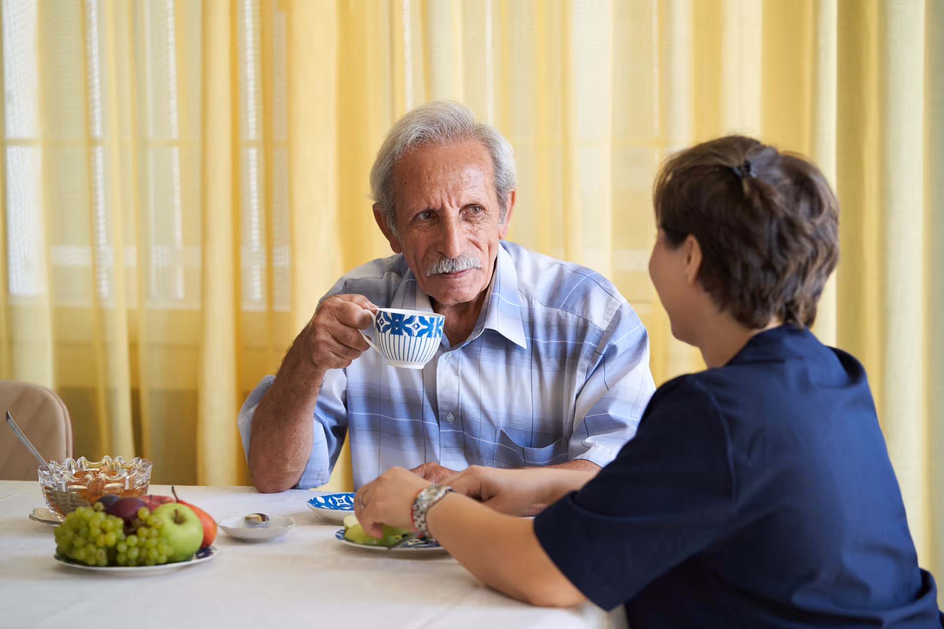 Elderly man sips from a patterned cup as caregiver chats at a dining table.