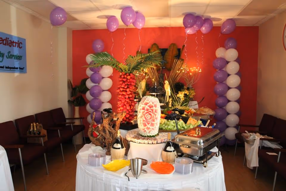 Indoor room decorated with purple balloons and a central fruit and snack buffet.