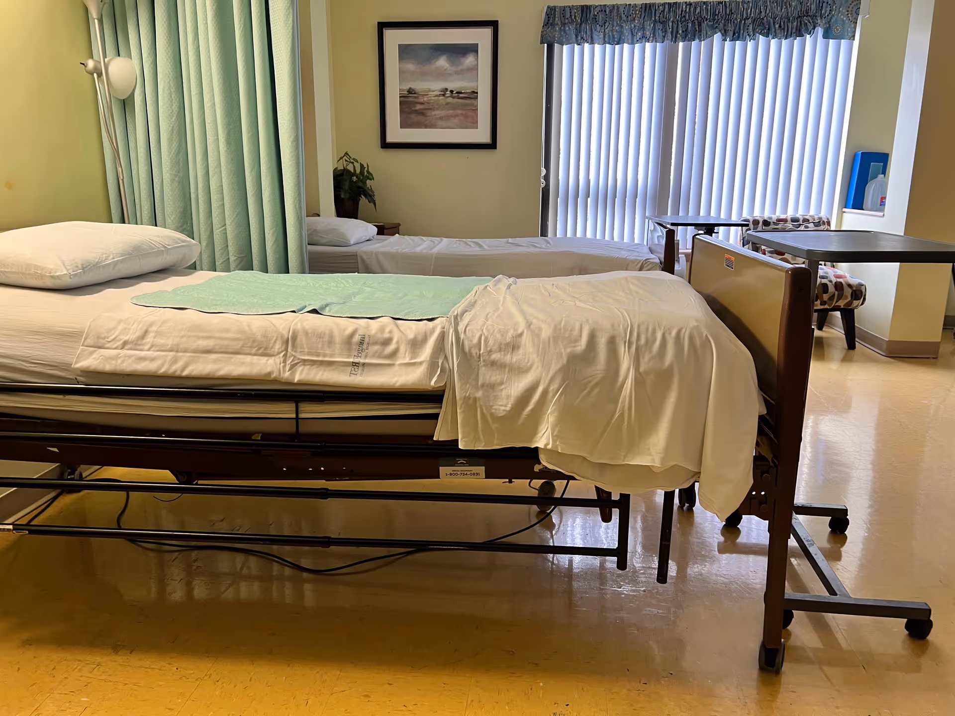 Two hospital-style beds beside privacy curtains in a bright patient room with blinds and a table.