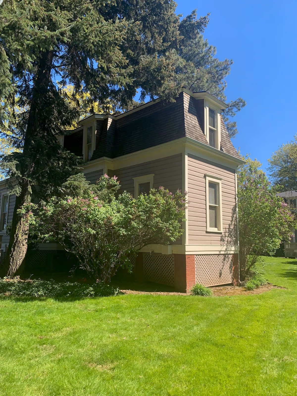 Beige two-story house with dormer windows, bushes, and a green lawn under a blue sky.