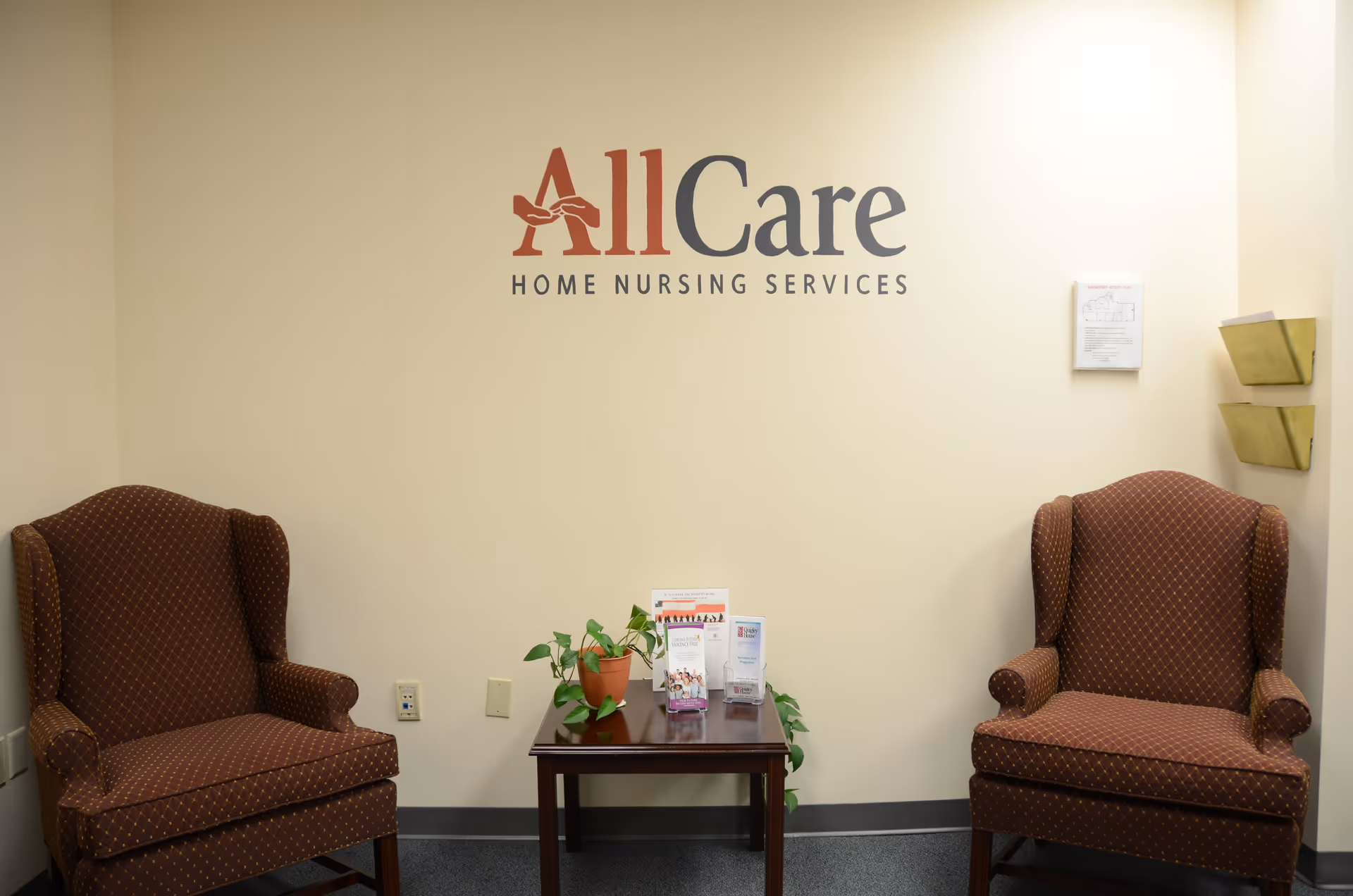 Waiting area with two burgundy chairs, a small table, plants, brochures, and AllCare signage on wall.