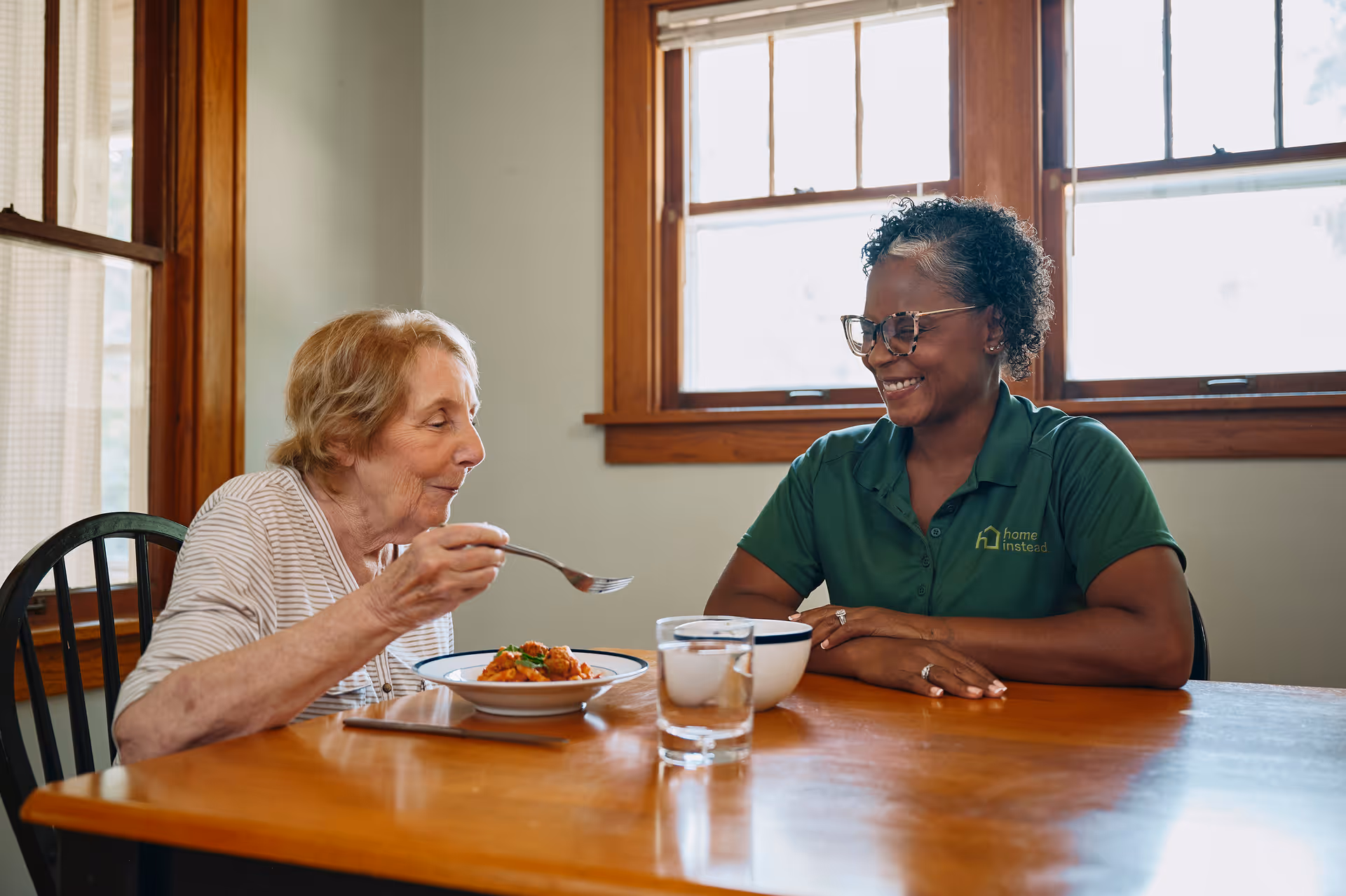 An elderly woman eats at a wooden dining table with a smiling caregiver.