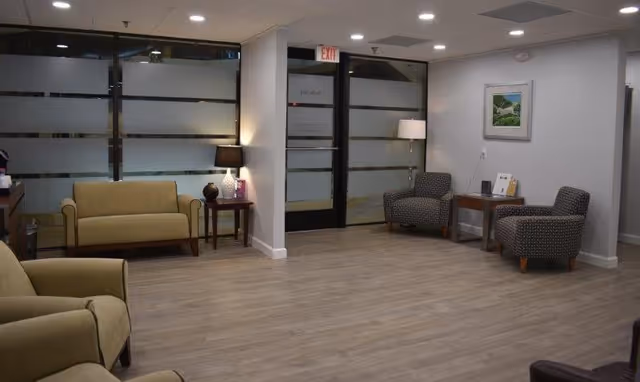 A spacious senior living lounge with beige seating, chairs, tables, lamps, and frosted glass partitions.