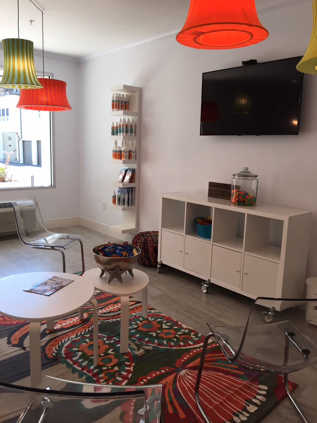 Bright modern lounge with colorful pendant lamps, glass chairs, shelving, rug, and a wall-mounted TV.