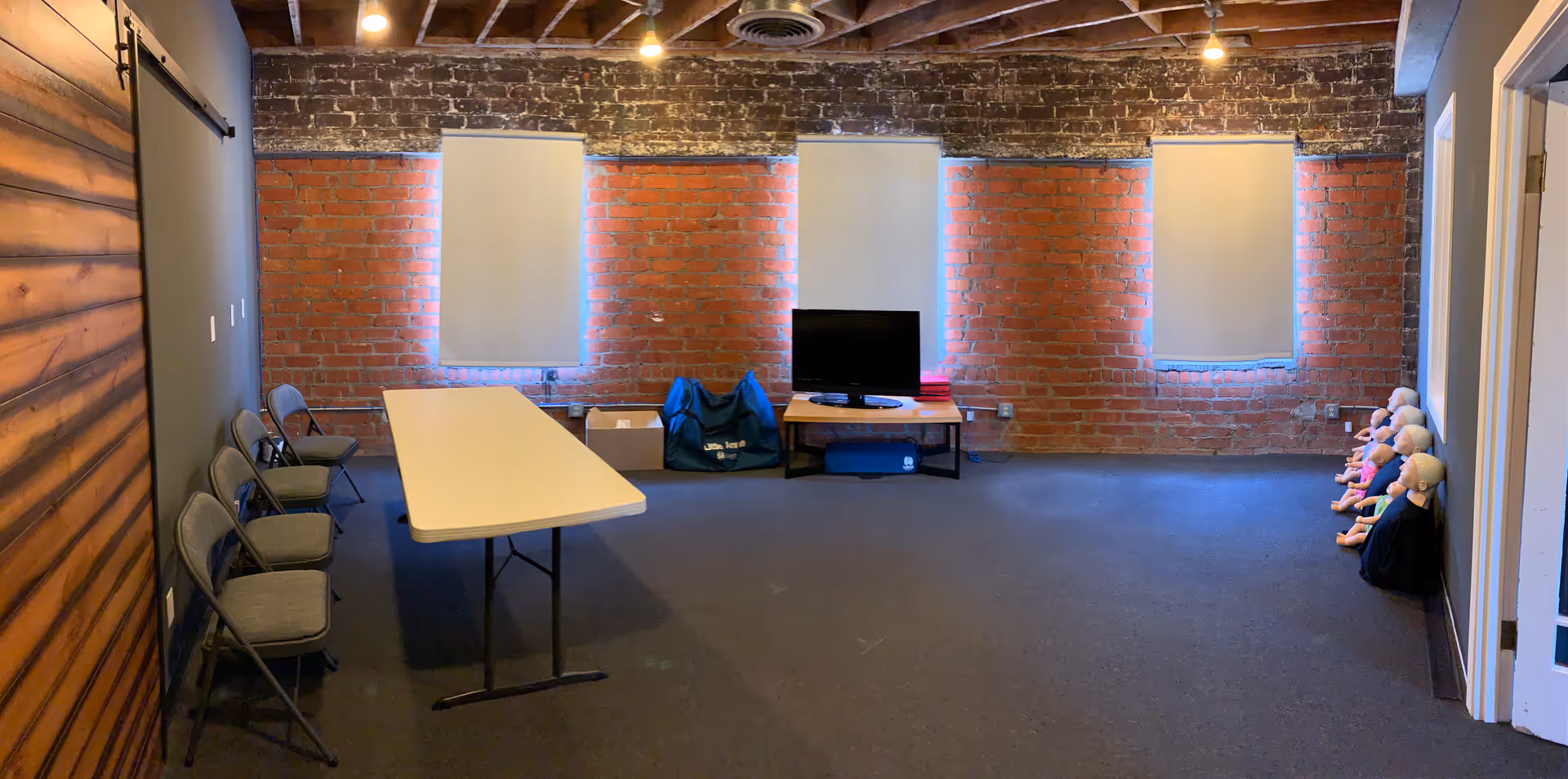 A brick-walled CPR training room with a long table, TV, and mannequin dolls along the right.