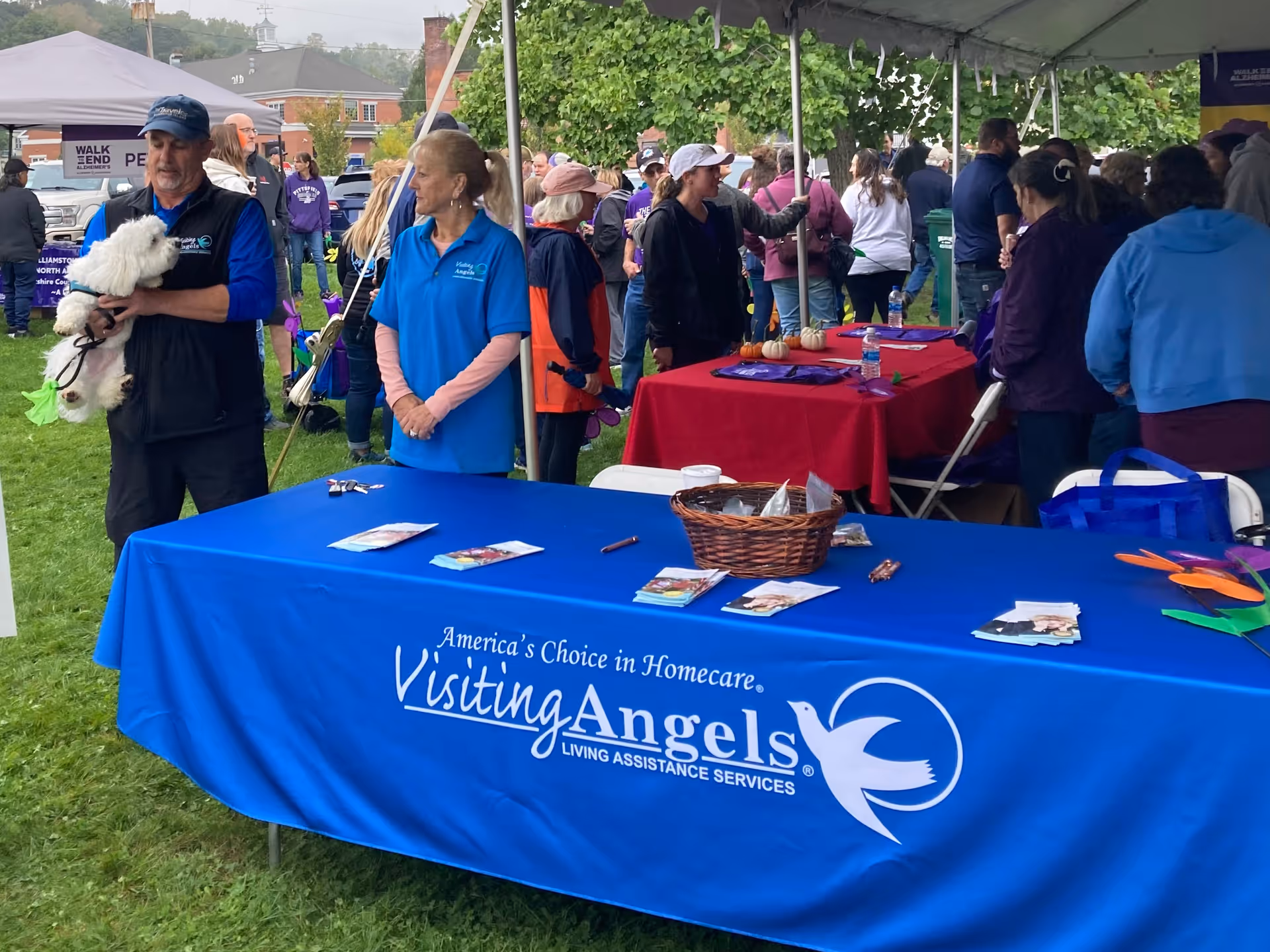 Outdoor community event with a Visiting Angels booth under tents on a grassy lawn.