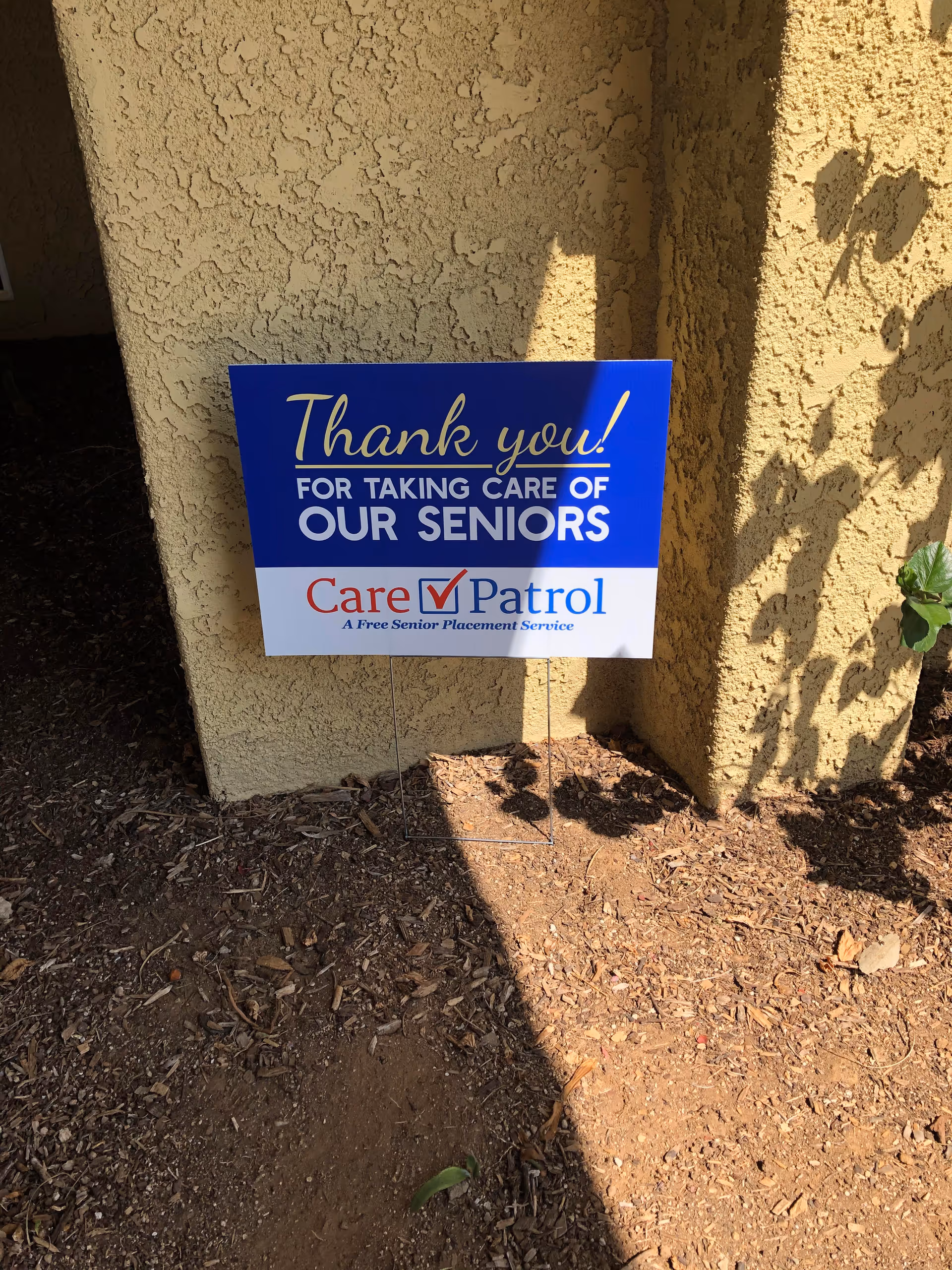 Blue and white sign thanking caregivers staked beside dirt by a stucco wall.