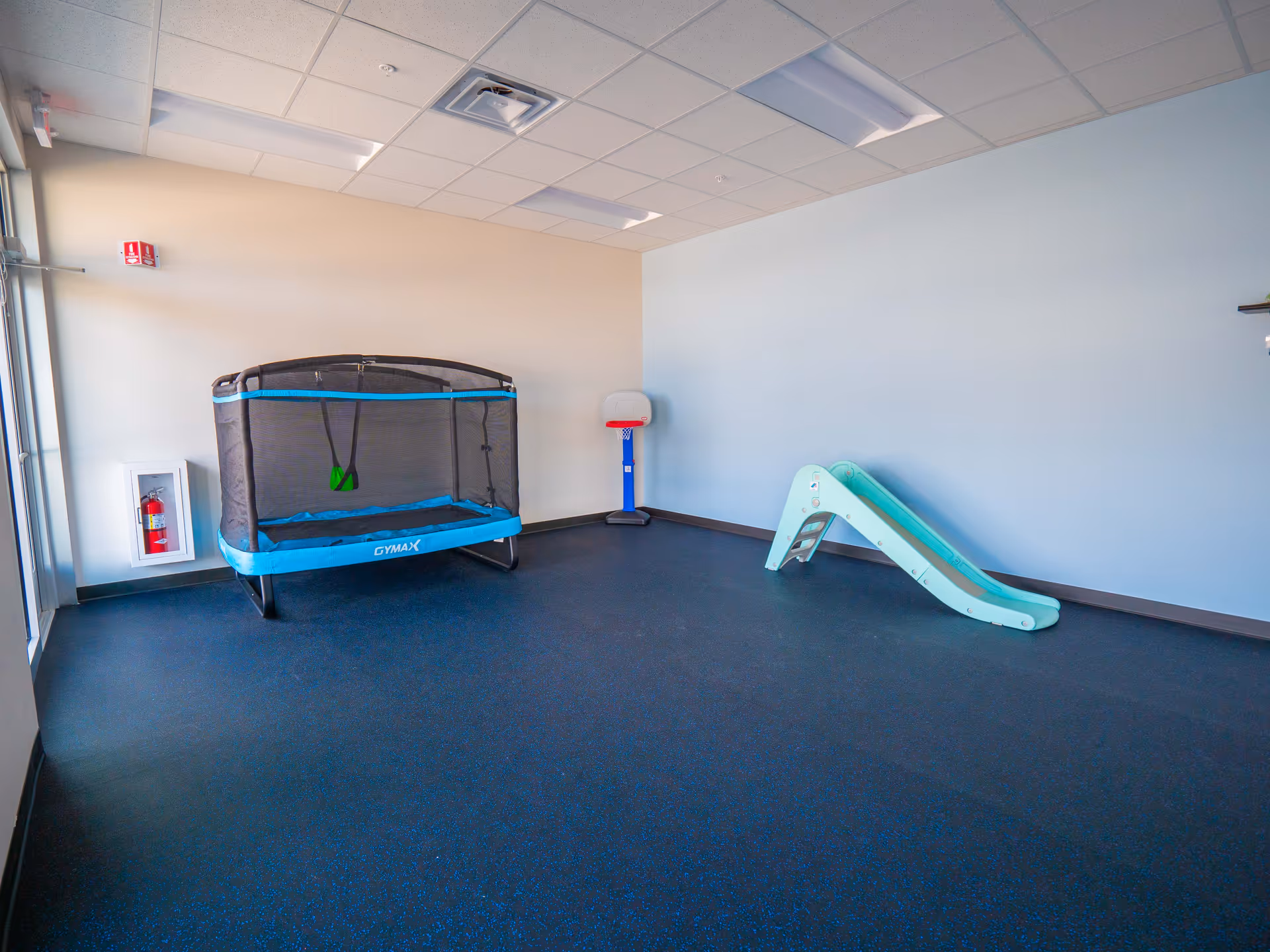 Indoor playroom with a trampoline, small slide, and a basketball hoop against light walls.
