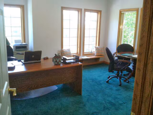 Office with wooden desk, laptop, printer, chairs, and large windows in a teal carpeted room.