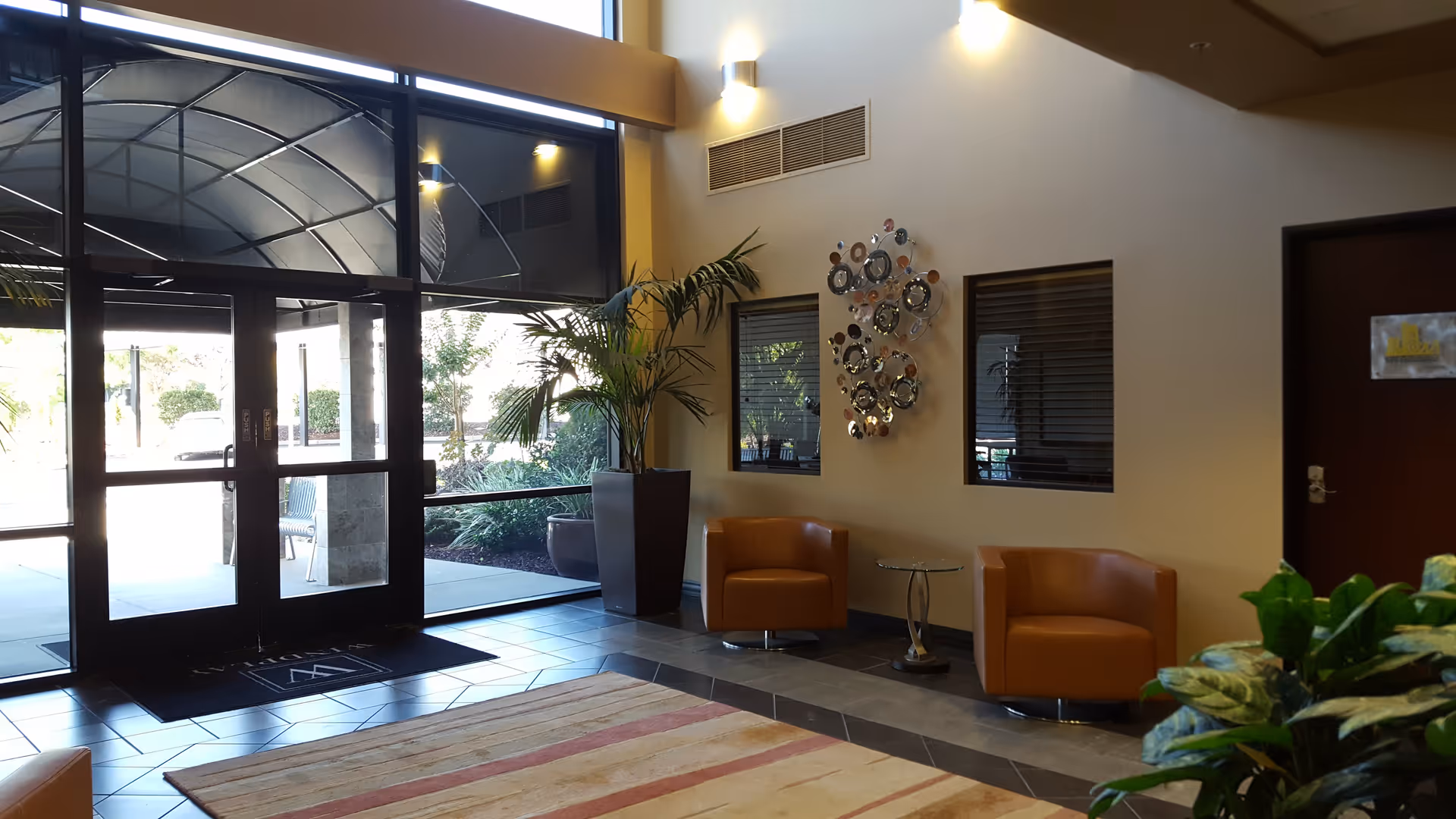 A modern senior living facility lobby with glass entry doors, orange chairs, plants, and wall art.