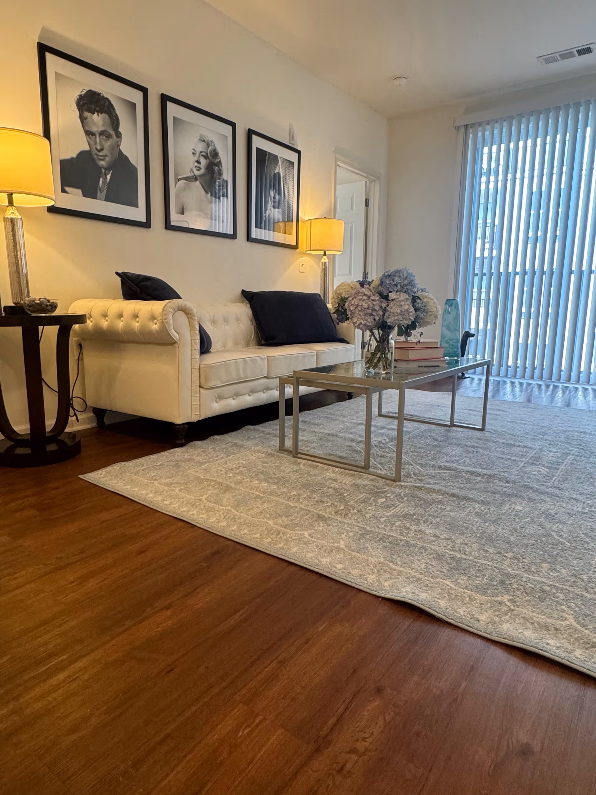 Bright living room with white tufted sofa, framed portraits, lamps, and a glass coffee table.