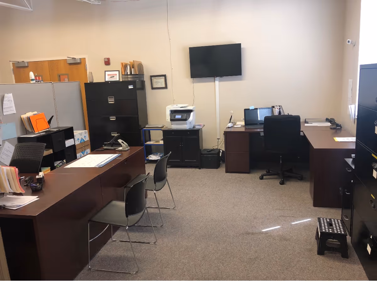 Office workspace with desks, chairs, filing cabinets, and a wall-mounted screen.