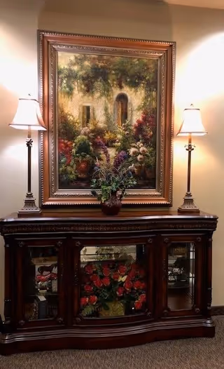 Dark wood cabinet with glass doors, framed painting above, two lamps, and a potted plant.
