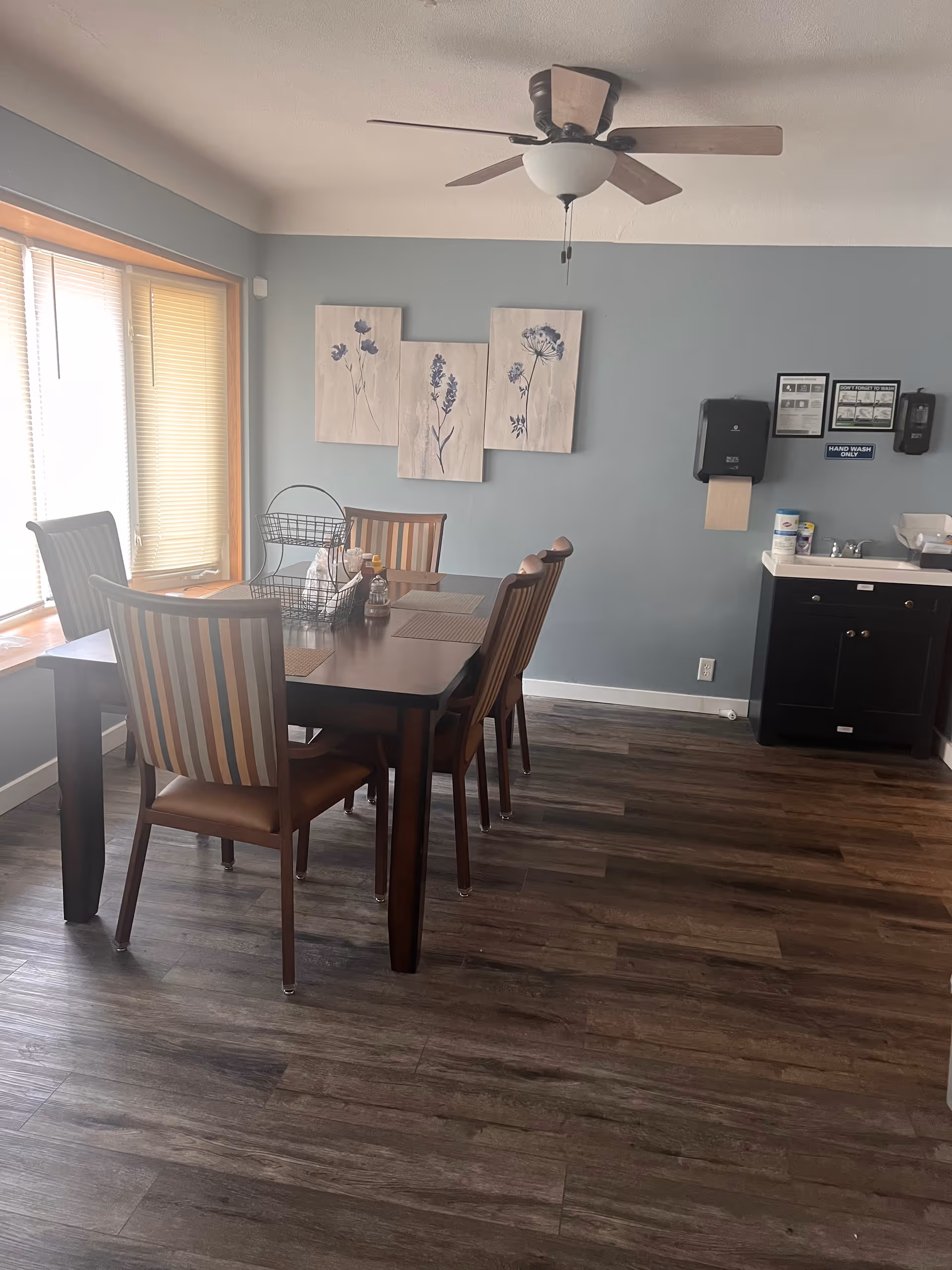 Dining room in a care facility with a table, chairs, wall art, and a ceiling fan.