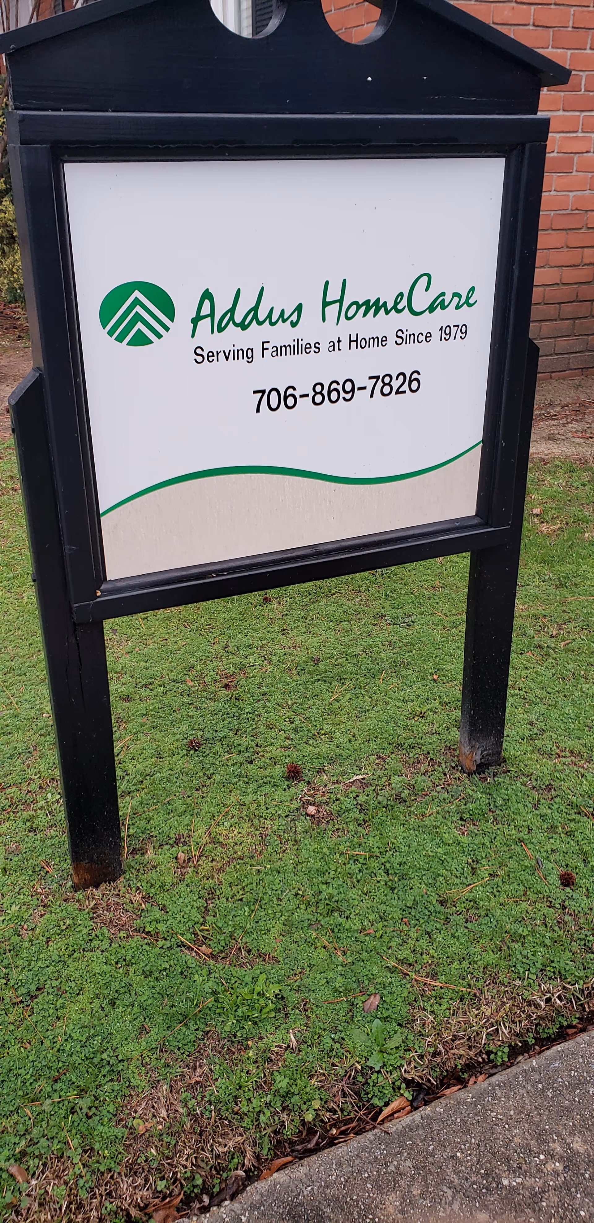 Outdoor sign for Addus HomeCare in a black frame by a brick building, with phone number.
