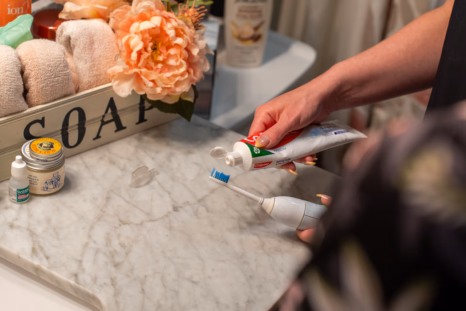 Person's hand squeezes toothpaste onto an electric toothbrush at a marble bathroom counter.