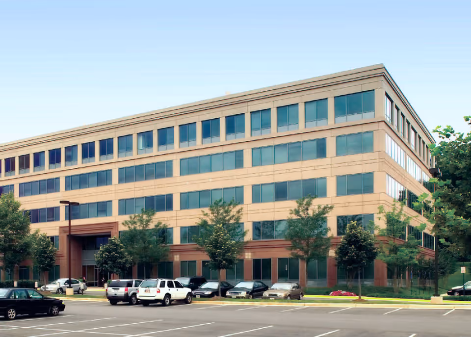 Exterior view of a large multi-story building with windows, parking, and trees.