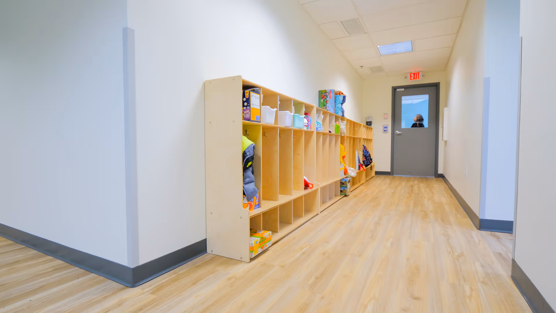 Long hallway with wooden floor, left cubby shelving, and an exit door at the far end.