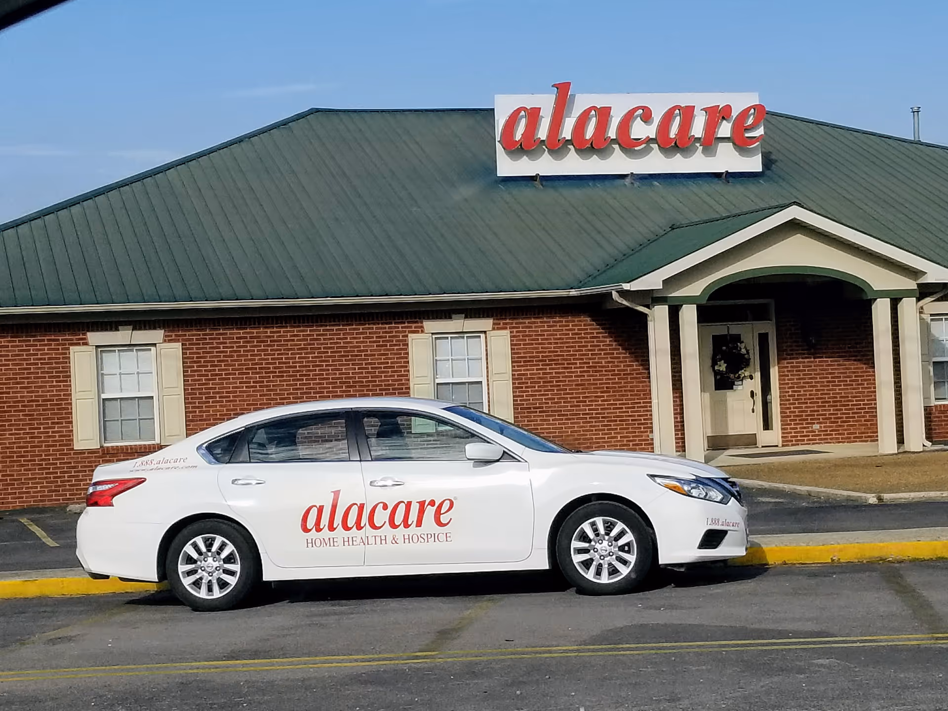Exterior of a brick facility with a green roof and a white Alacare car parked front.