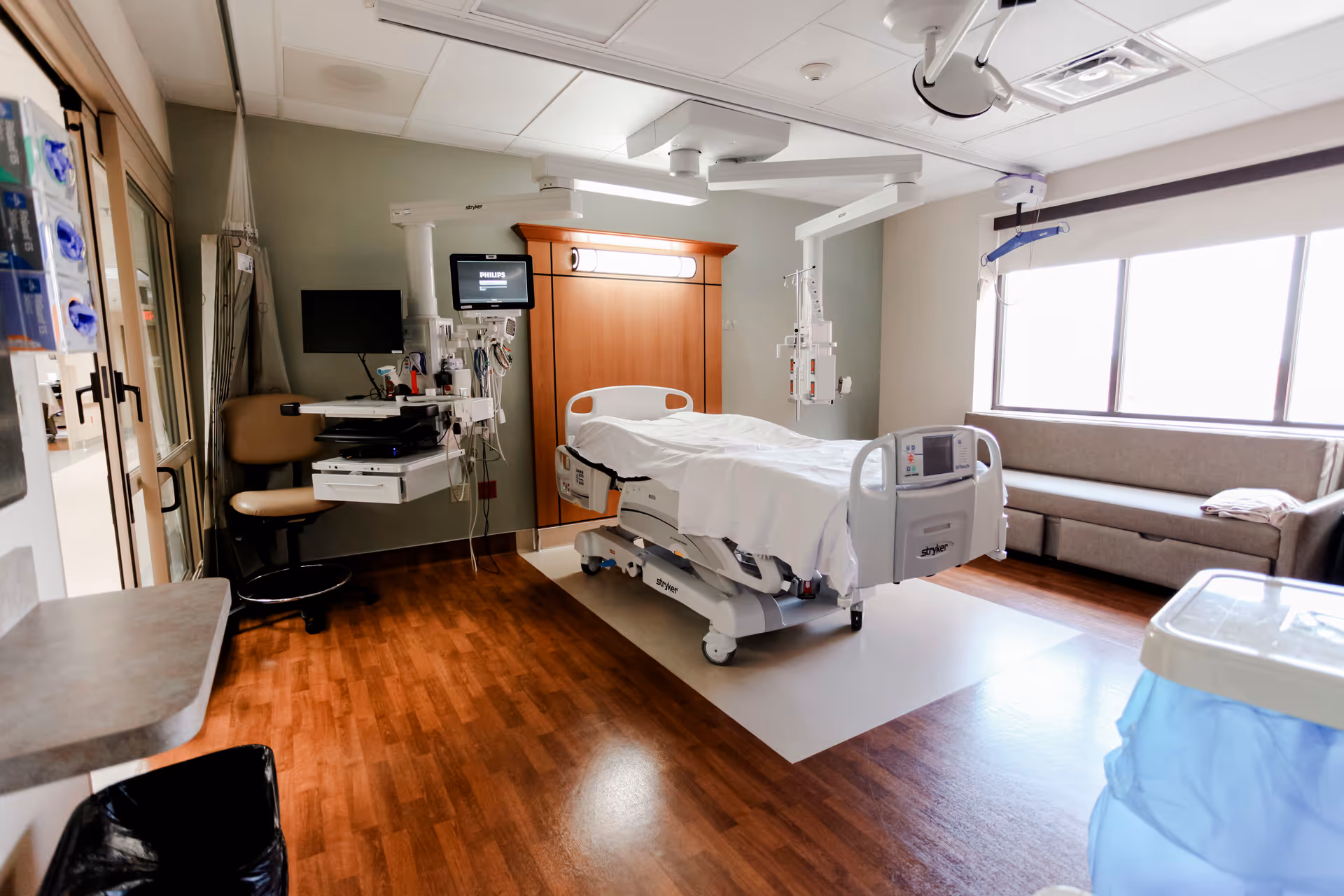 Hospital room with a patient bed, monitors, IV pole, and a seating area by large windows.